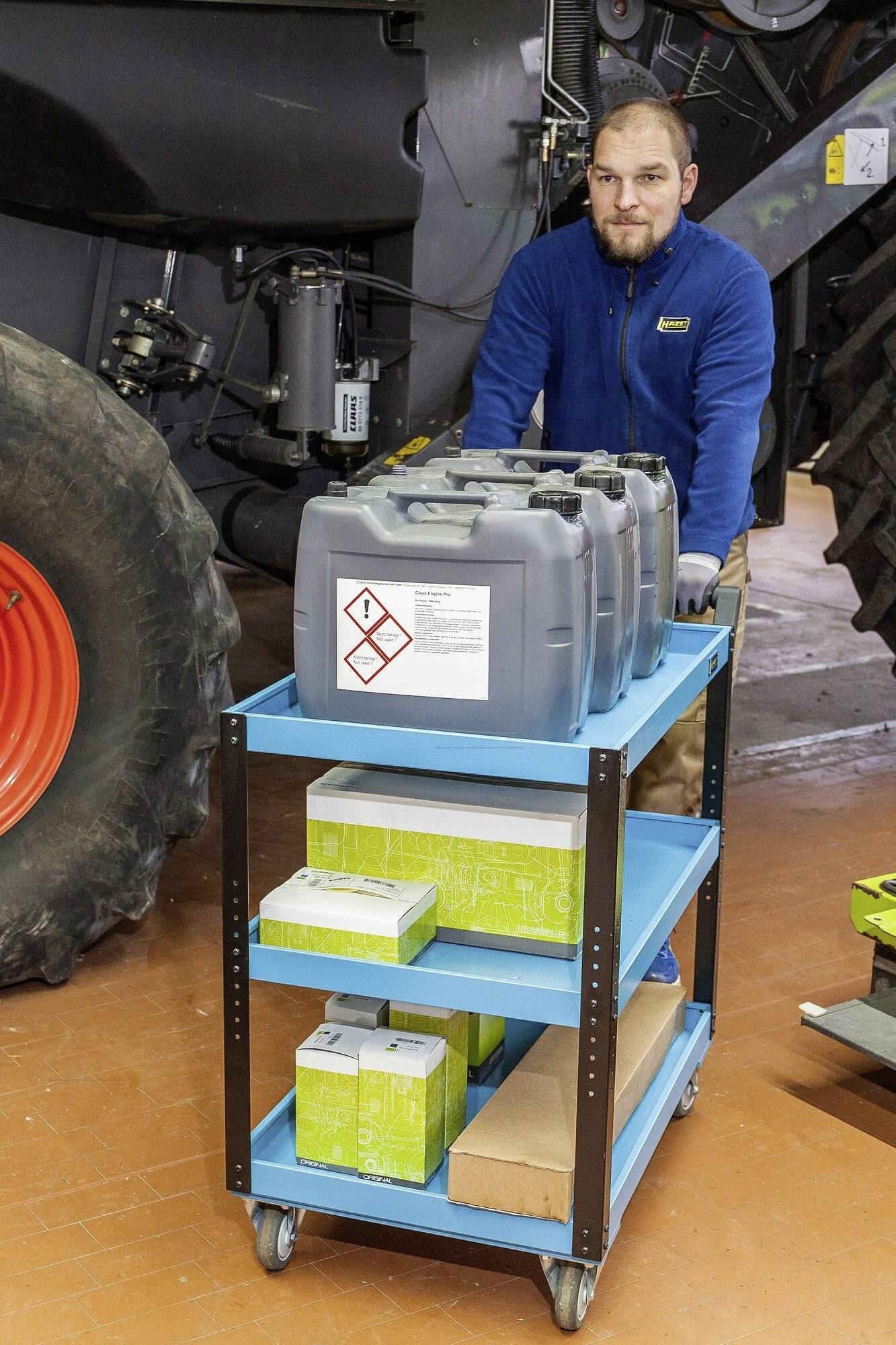 Ein Mann schiebt einen blauen Rollwagen mit Benzinkanistern neben großen Landmaschinen.