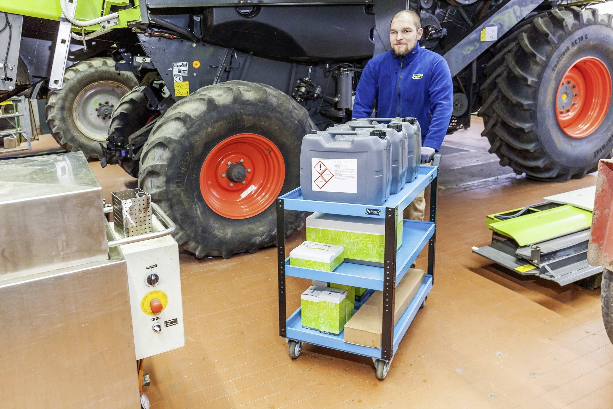 Ein Mann schiebt einen Wagen mit Kanistern und Kisten in einer Werkstatt. Im Hintergrund steht ein großes landwirtschaftliches Fahrzeug.