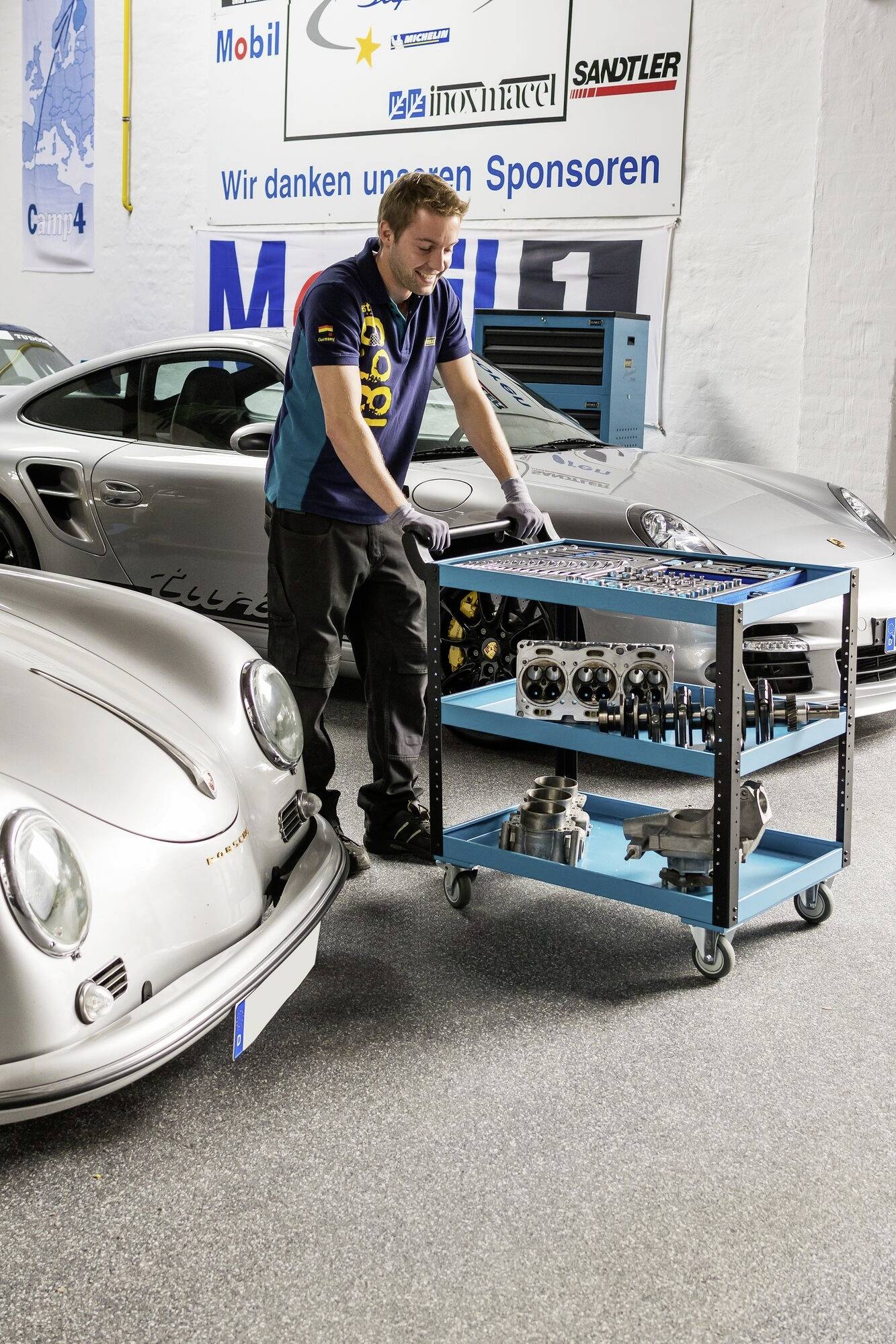 Ein Mechaniker steht zwischen zwei Sportwagen in einer Werkstatt; er arbeitet an einem Werkzeugwagen voller Autoteile.