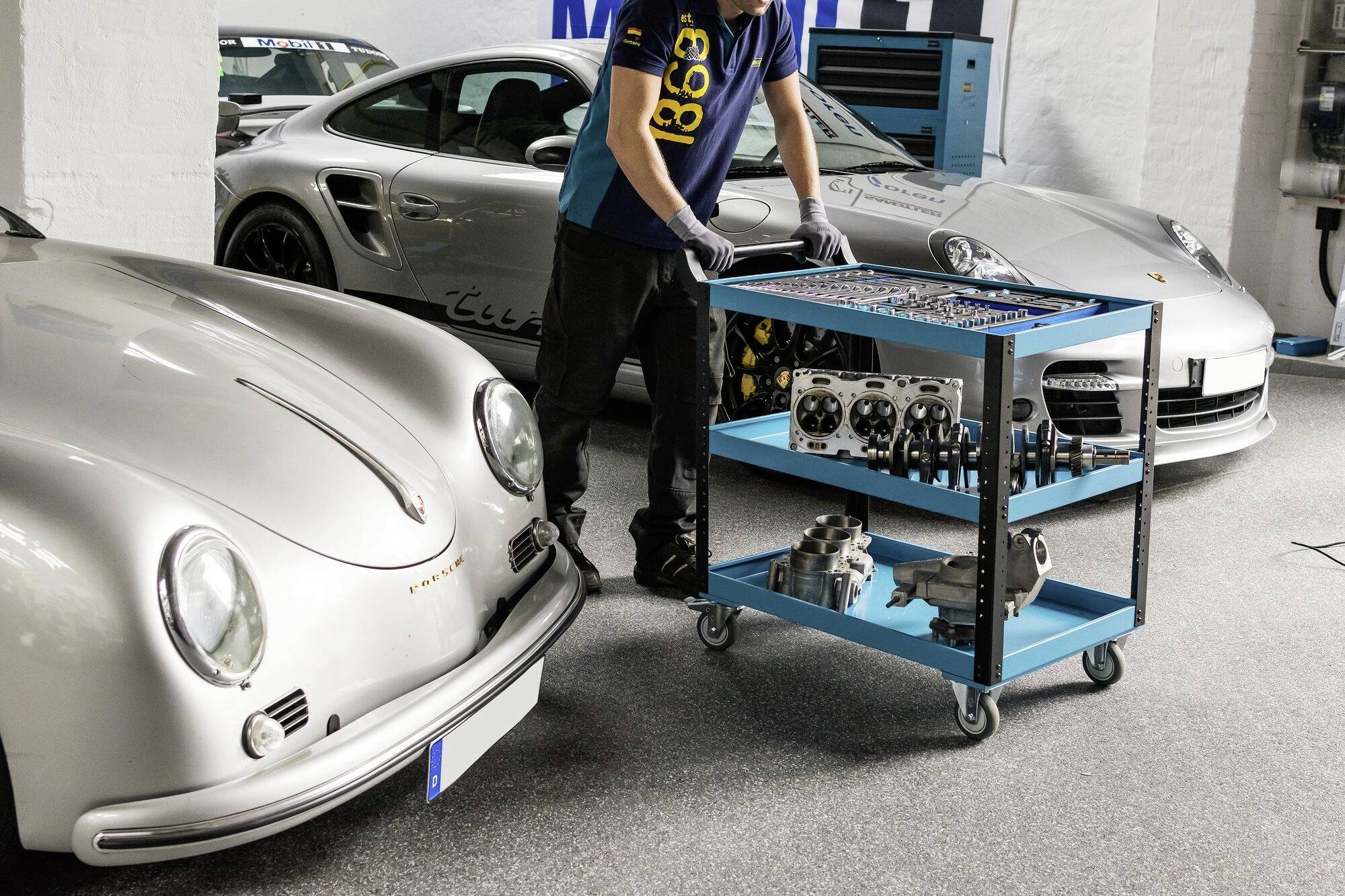 'Person in Werkstatt' bewegt einen Werkzeugwagen zwischen zwei silbernen Sportwagen. Enthält verschiedene Autoteile auf dem Wagen.