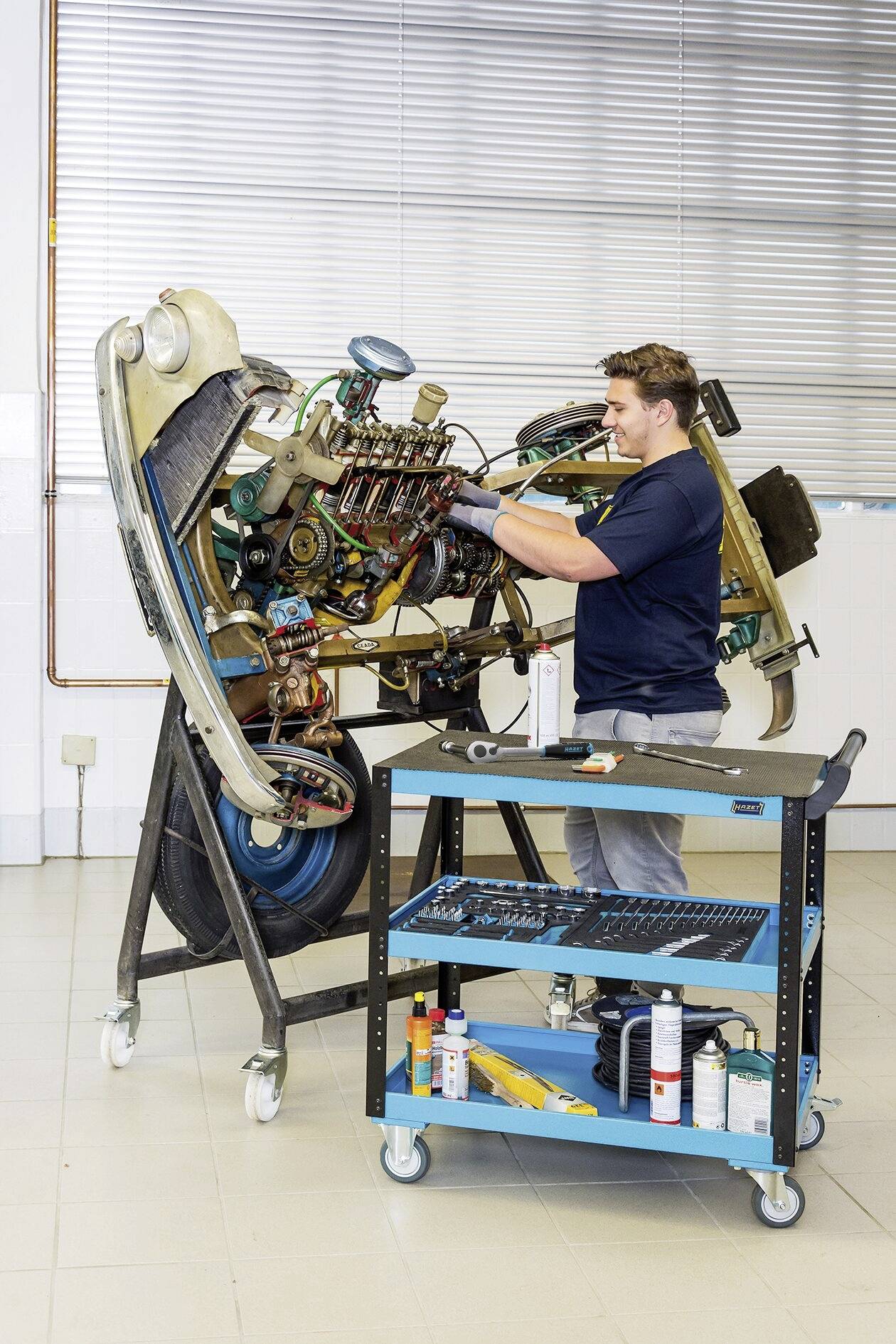 Ein Techniker arbeitet an einem montierten Flugzeugtriebwerk in einer Werkstatt. Ein blauer Werkzeugwagen steht daneben.