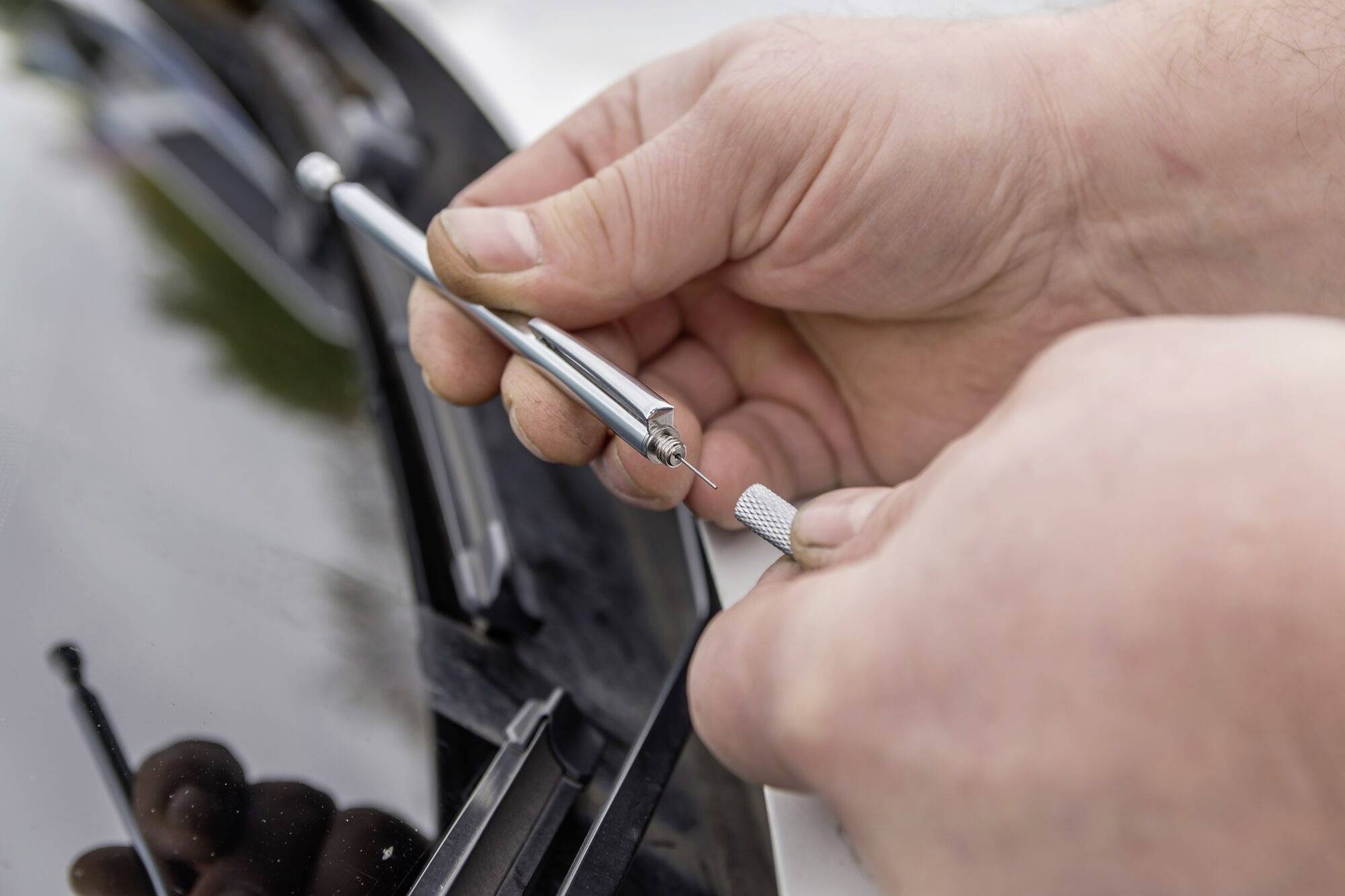 Nahaufnahme einer Person, die ein kleines Werkzeug benutzt, um einen Steinschlag auf einer Autoscheibe zu reparieren.