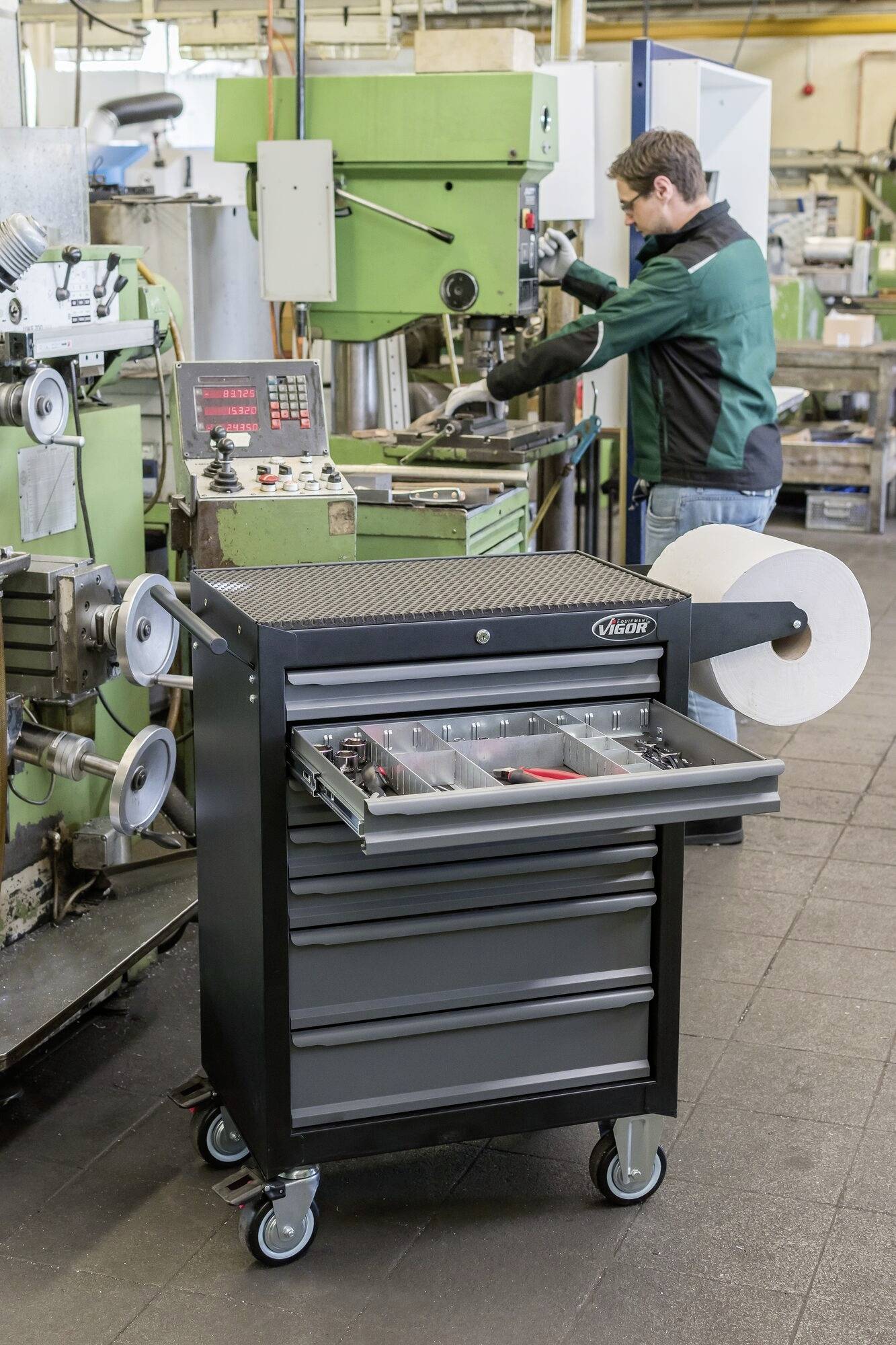 Ein Mann arbeitet in einer Werkstatt an einer Maschine. Im Vordergrund steht ein Werkzeugwagen mit Schubladen und Papierrollenhalter.