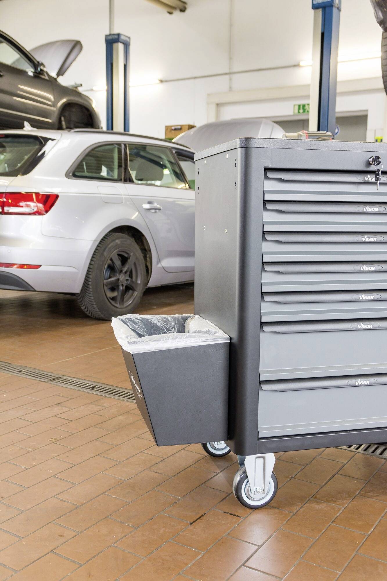 In einer Autowerkstatt steht ein silbernes Auto auf einer Hebebühne. Im Vordergrund befindet sich ein grauer Werkzeugwagen mit sechs Schubladen.