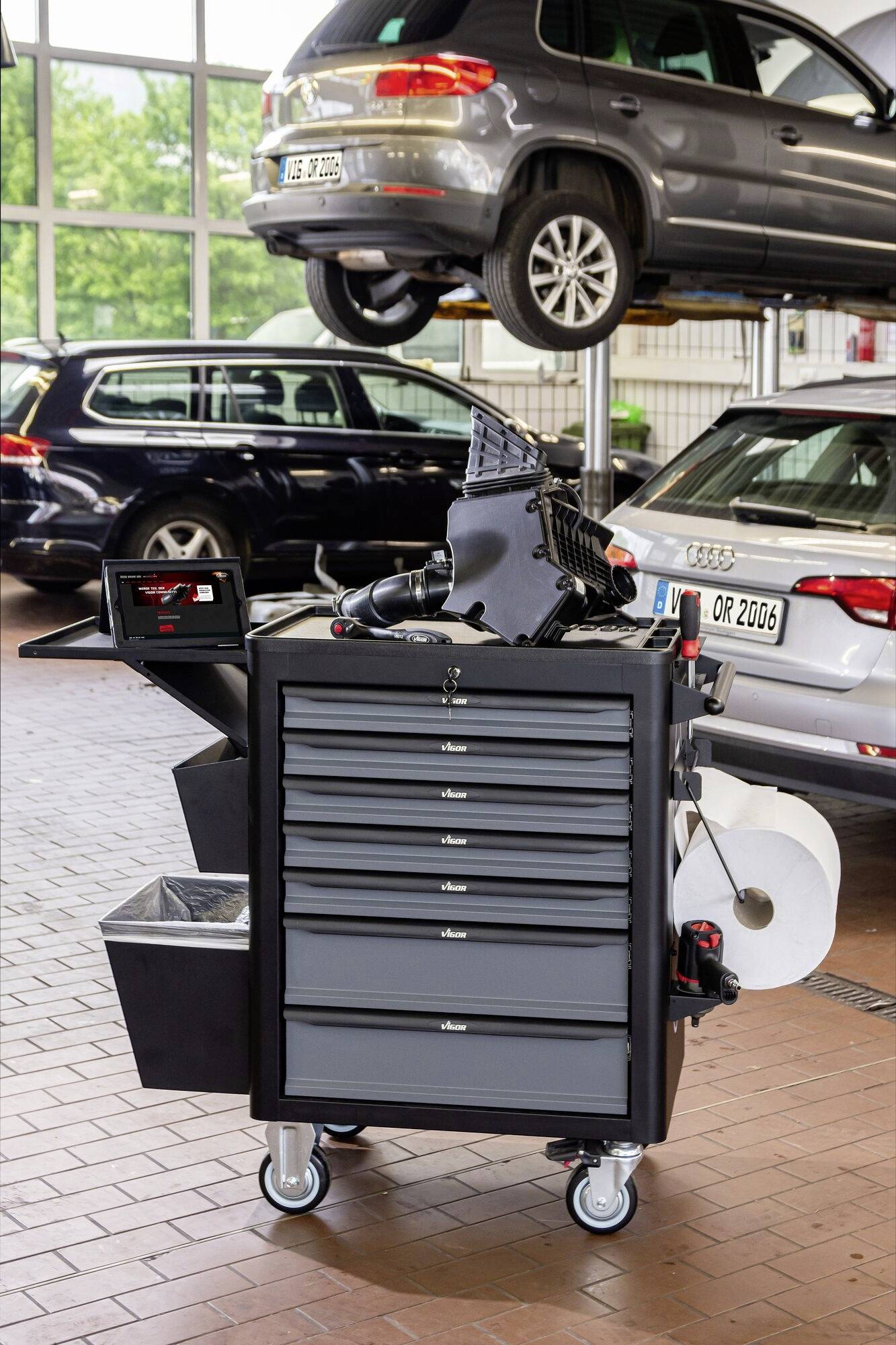 Ein Werkstattwagen steht in einer Autowerkstatt vor einem aufgebockten Auto. Er enthält Werkzeuge und Papierrollen für Reparaturen.