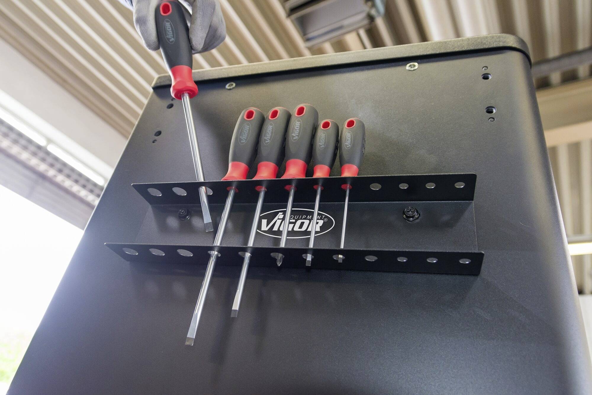 Ein Metallhalter mit sechs Schraubendrehern, montiert an einem Schrank. Ein Handschuh hält einen Schraubendreher.