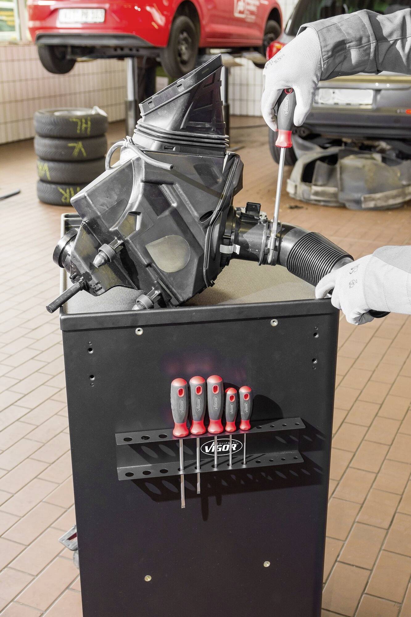 Ein Mechaniker arbeitet in einer Autowerkstatt an einem Fahrzeugteil auf einem Werkzeugwagen. Im Hintergrund sind Autoreifen zu sehen.