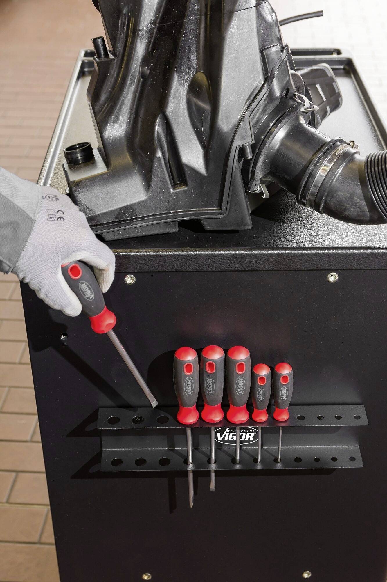 Eine Hand mit Handschuh greift nach einem Schraubendreher von einer auf einem Gerät montierten Halterung mit mehreren Schraubendrehern.