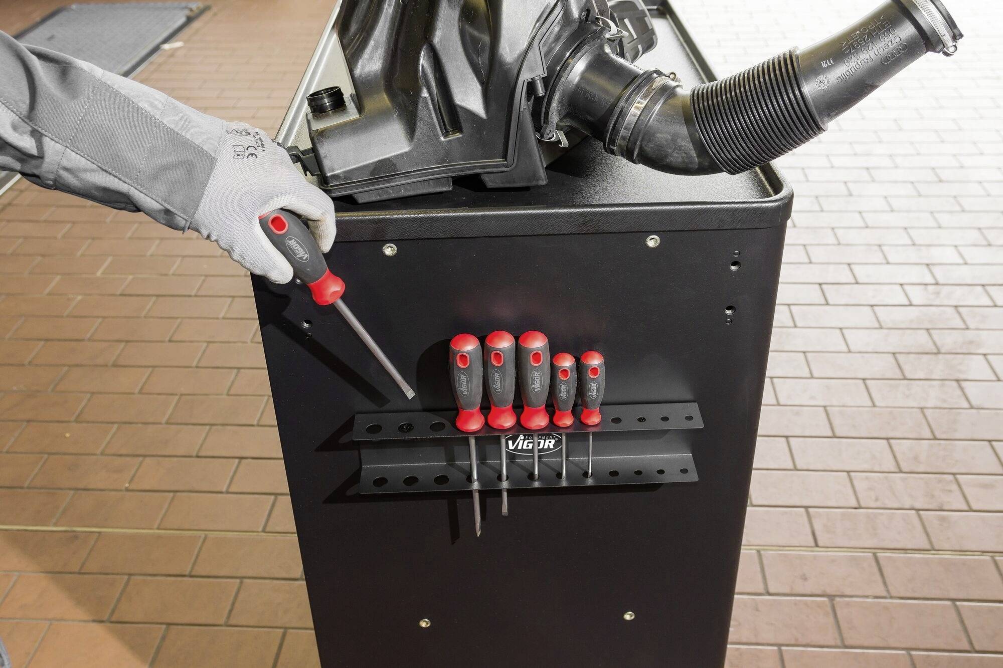 Eine Hand in einem grauen Handschuh greift nach einem Schraubenzieher an einem Werkzeugwagen mit mehreren Schraubenziehern vor einer Werkbank.