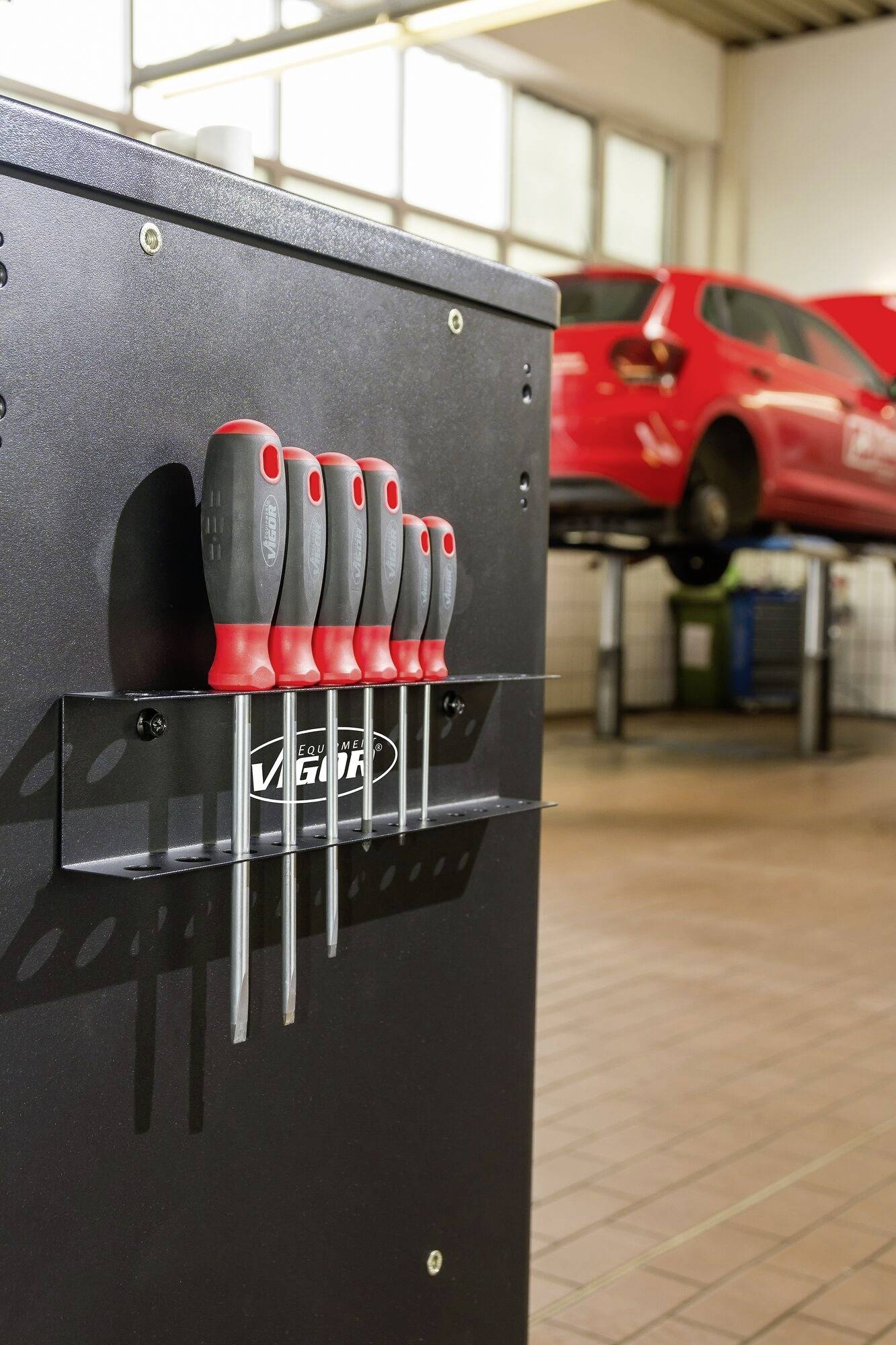 Ein Schraubendreher-Set hängt an einer Werkbank in einer Autowerkstatt, im Hintergrund steht ein rotes Auto auf einer Hebebühne.