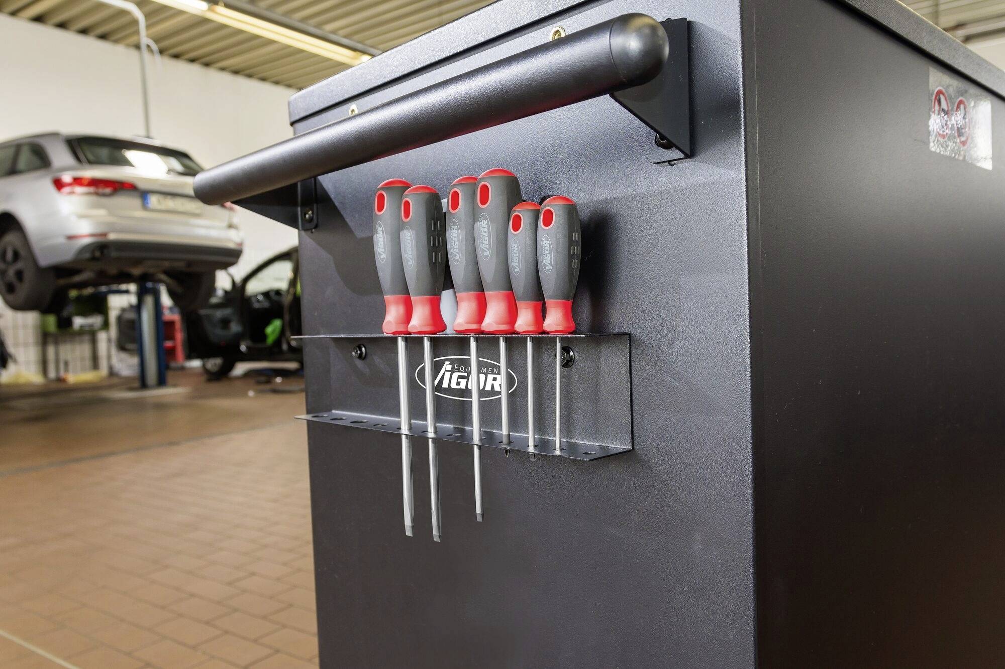 Ein Werkstattwagen mit rotem Griffschraubendrehern auf einem Werkzeughalter. Im Hintergrund ein angehobenes Auto auf einer Hebebühne.