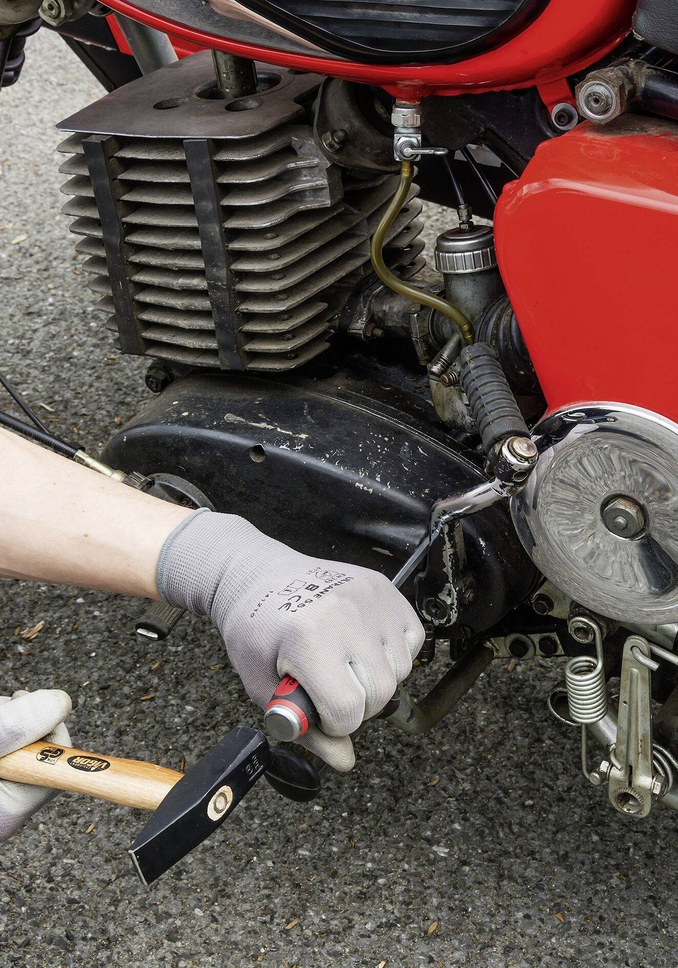 Eine Person trägt Schutzhandschuhe und repariert den Motor eines roten Motorrads mit einem Hammer auf einer asphaltierten Straße.