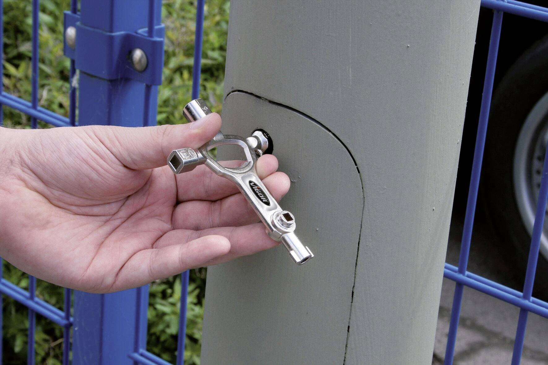 Eine Hand hält ein Ventilschlüssel, der an einem Schließmechanismus eines grauen Pfeilers verwendet wird, nahe einem blauen Metallzaun.