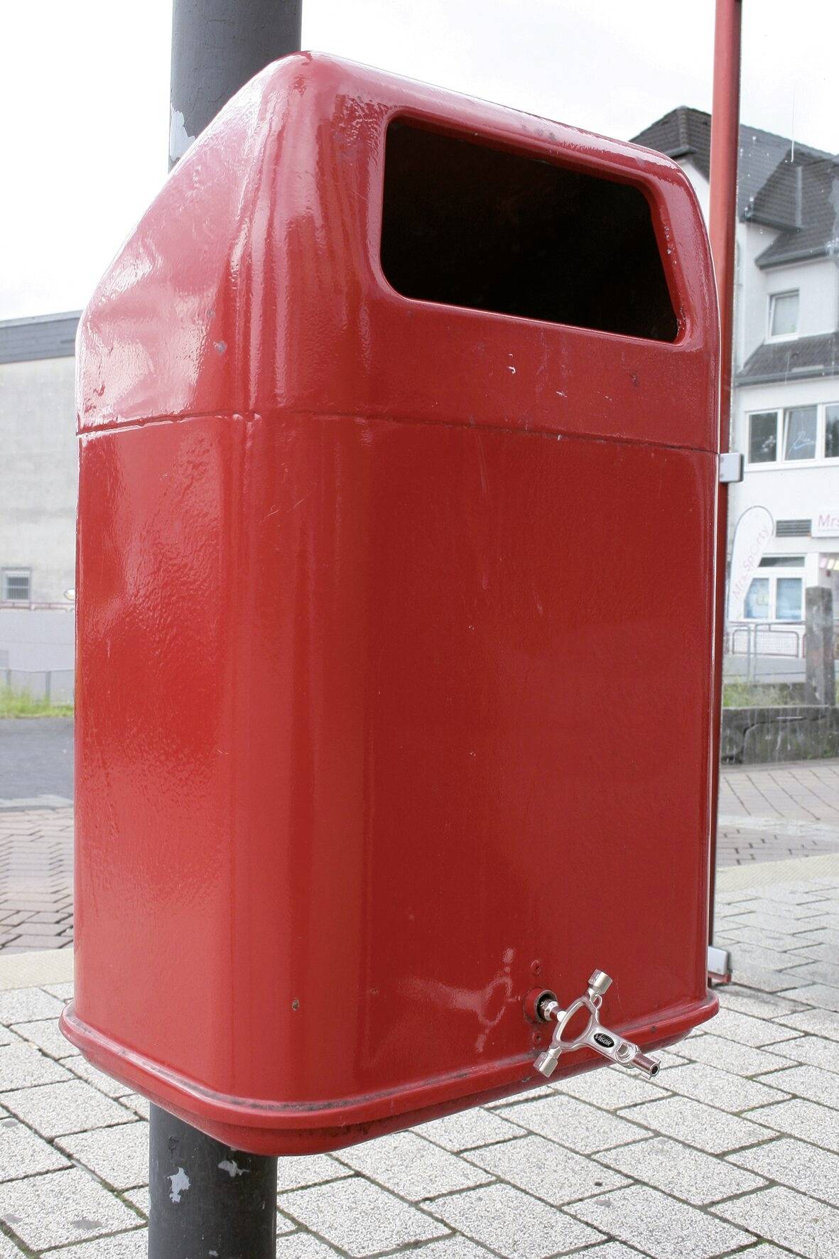 Roter Mülleimer an einer Straße, montiert an einem Metallpfosten. Im Hintergrund sind Gebäude zu sehen.