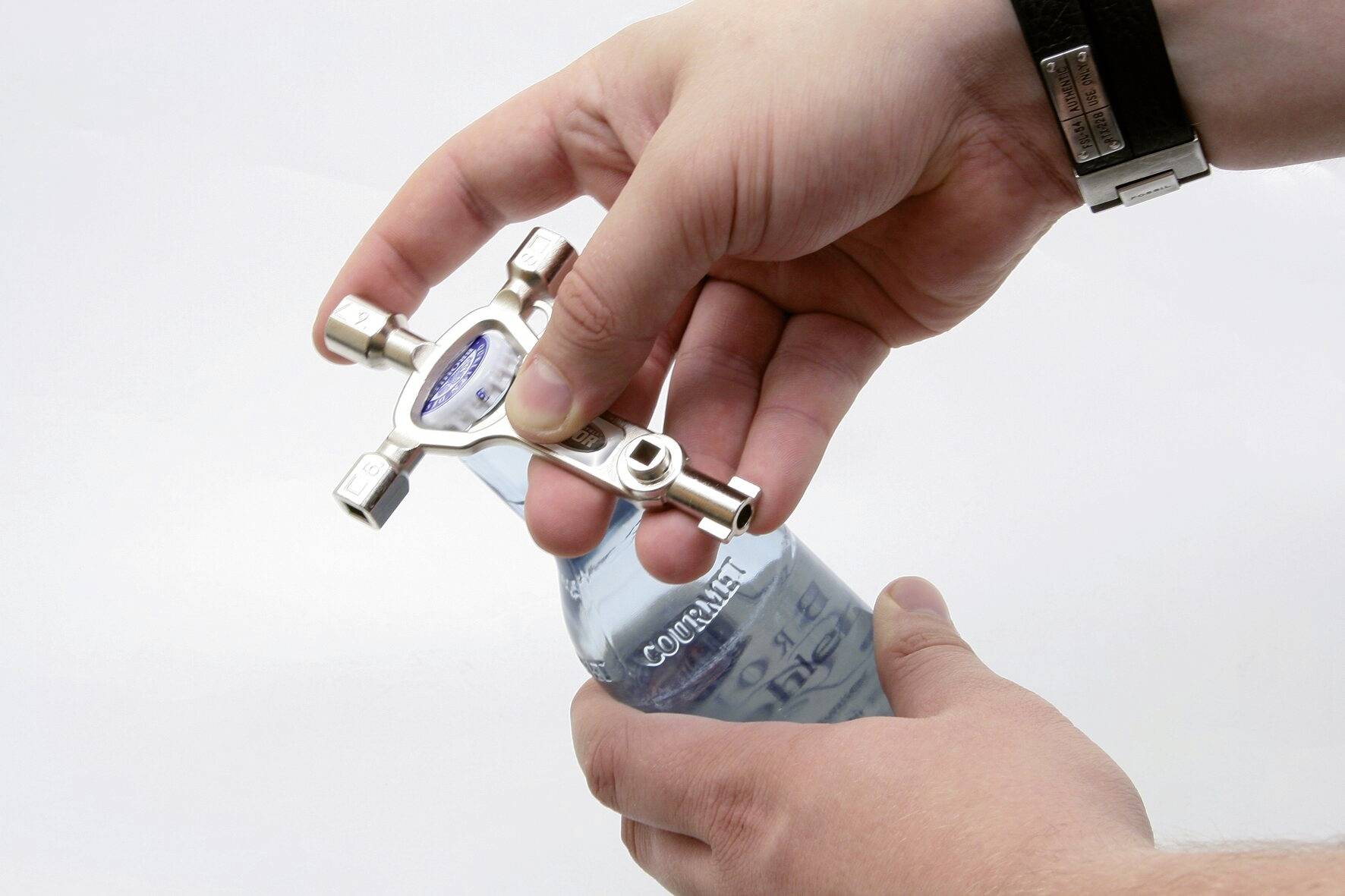 Zwei Hände öffnen eine Flasche mit einem silbernen Kronkorkenöffner. Die Flasche ist blau mit weißem Etikett.