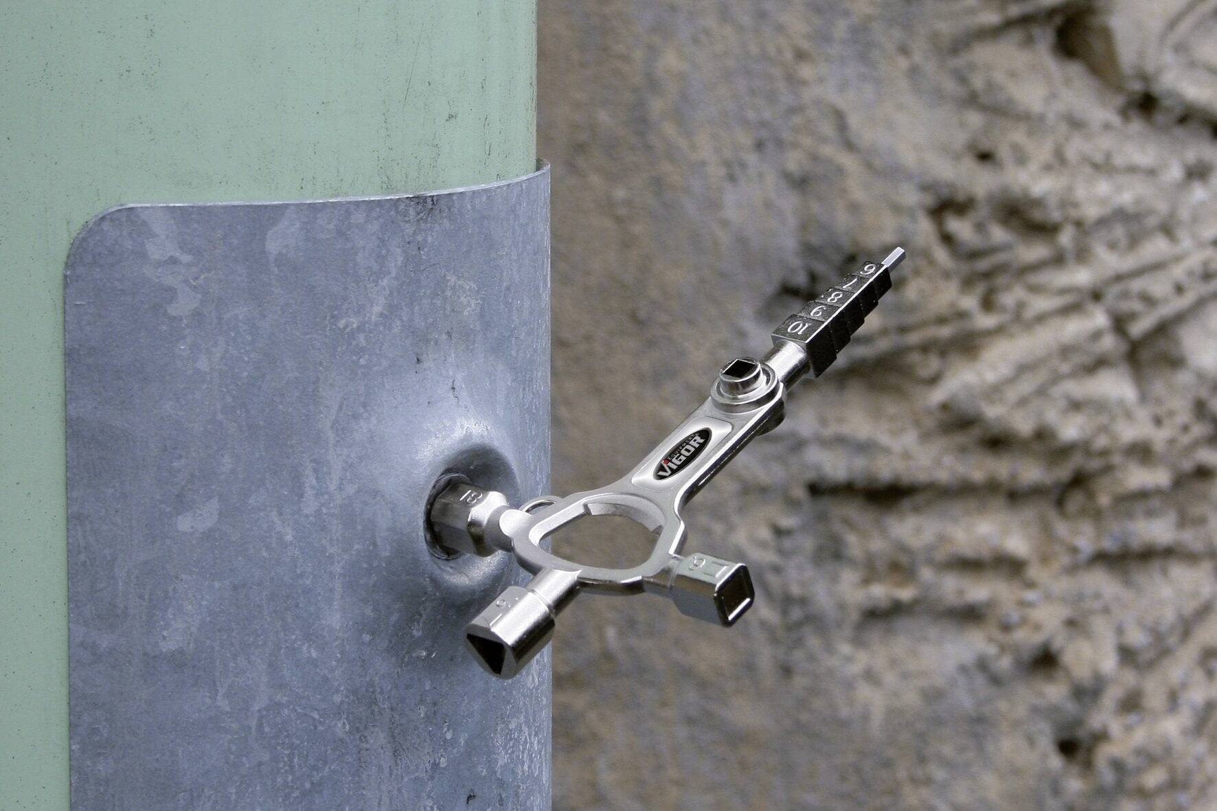 Ein silberner Universalschlüssel steckt in einem Metallkasten an einer grünen runden Säule vor einer rauen Steinwand.