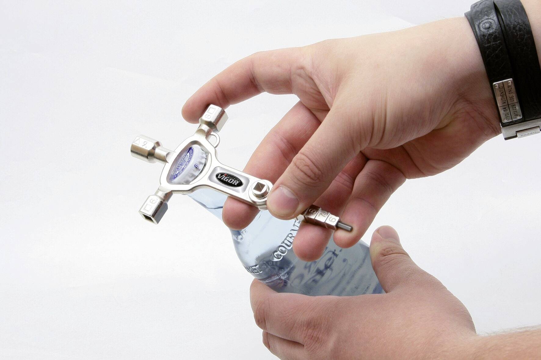 Eine Person hält ein Werkzeug mit mehreren Steckschlüsseln und öffnet damit eine Glasflasche, die teilweise sichtbar ist.