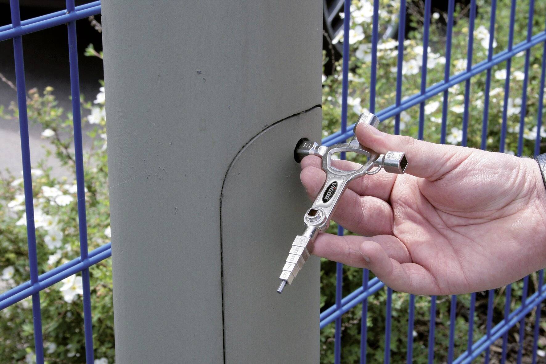 Eine Hand benutzt einen Vierkantschlüssel, um ein Schloss an einem Metallpfosten neben einem blauen Drahtzaun zu öffnen.