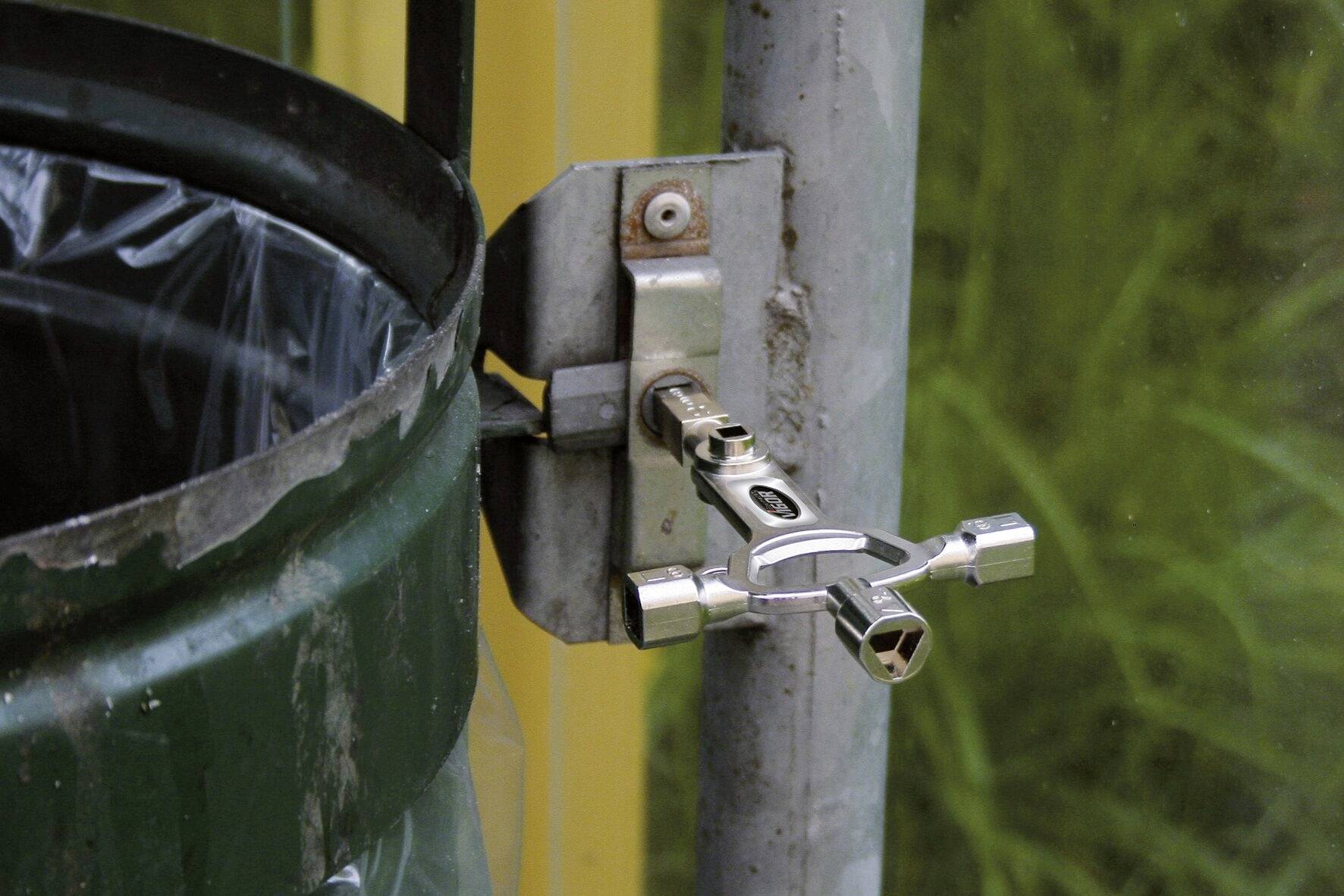 Ein universeller Schlüsselschlüssel steckt in einer Halterung an einer grünen Mülleimerabdeckung, offenbar bereit zur Nutzung.