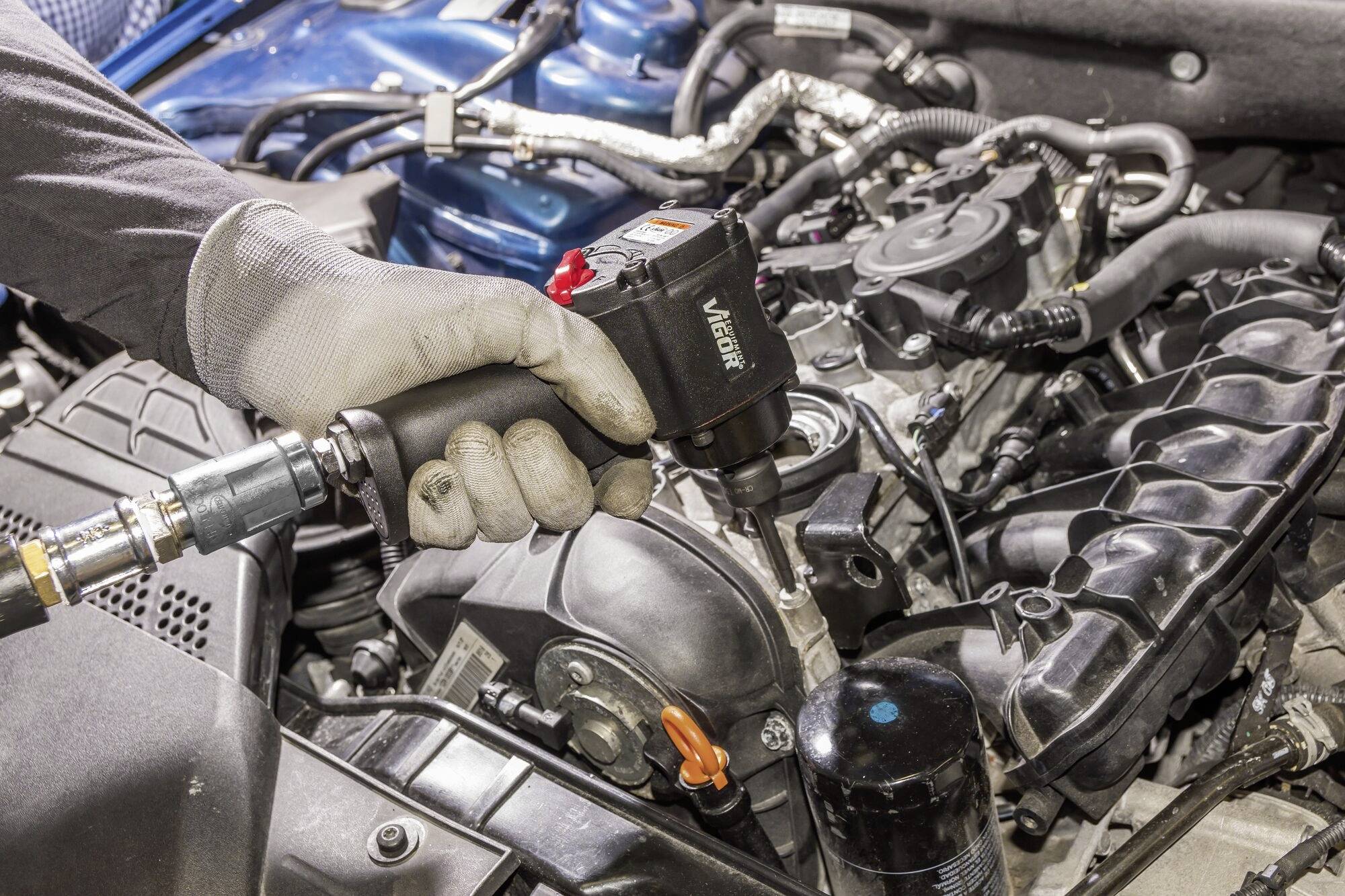 Eine Person mit Handschuhen verwendet ein pneumatisches Werkzeug, um an einem Autoteilmotor zu arbeiten. Verschiedene Motorkomponenten sind sichtbar.