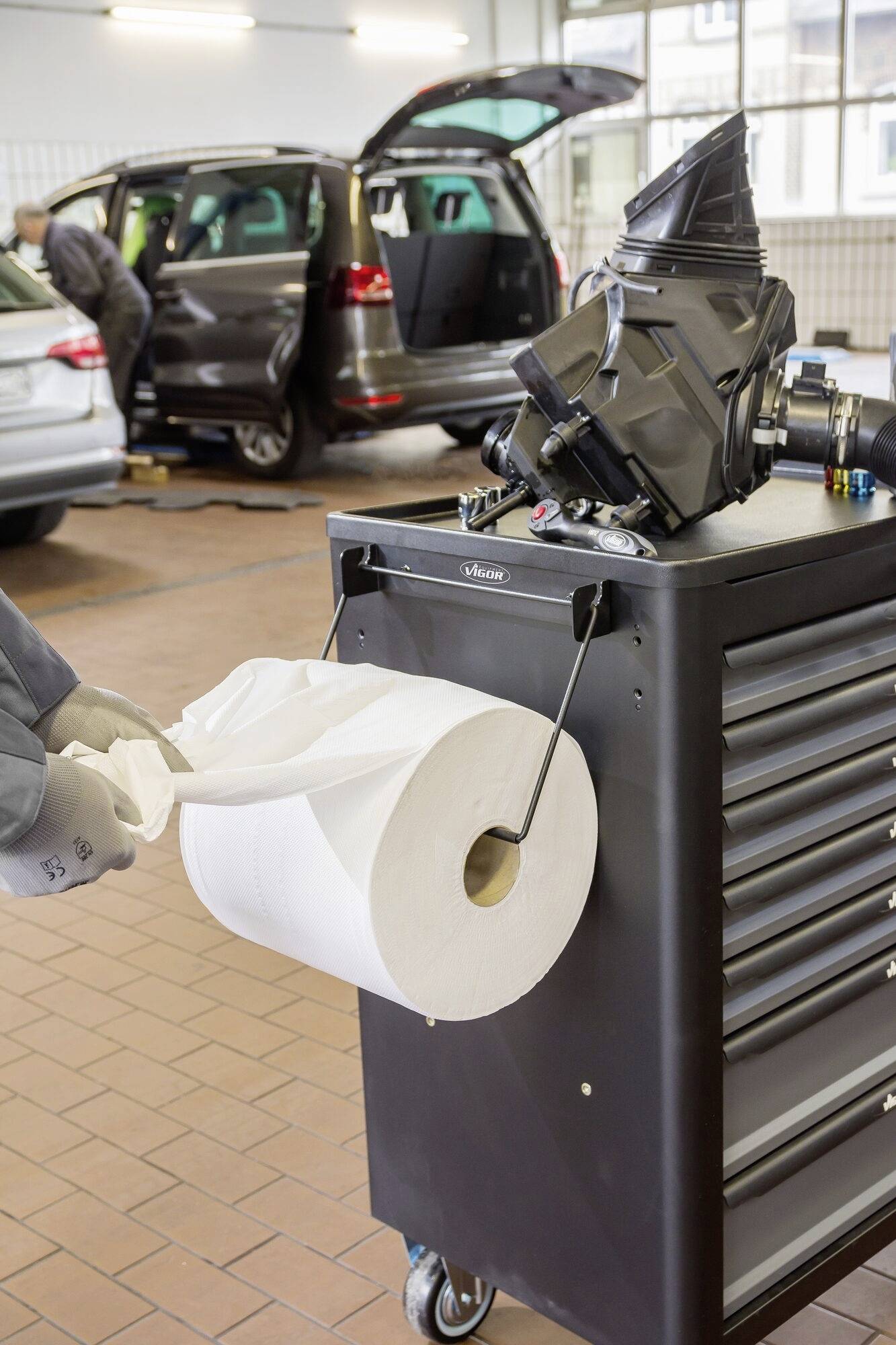 Ein Automechaniker nutzt Papiertücher an einem Werkzeugwagen in einer Kfz-Werkstatt. Im Hintergrund wird an einem Auto gearbeitet.