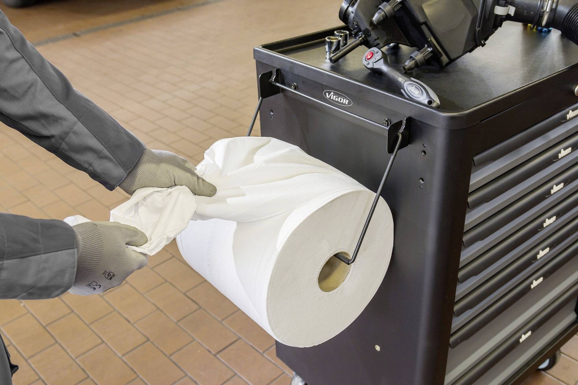 Eine Person zieht ein Papiertuch von einer großen Rolle, die an einem Werkstattwagen befestigt ist. Handschuhe werden getragen.