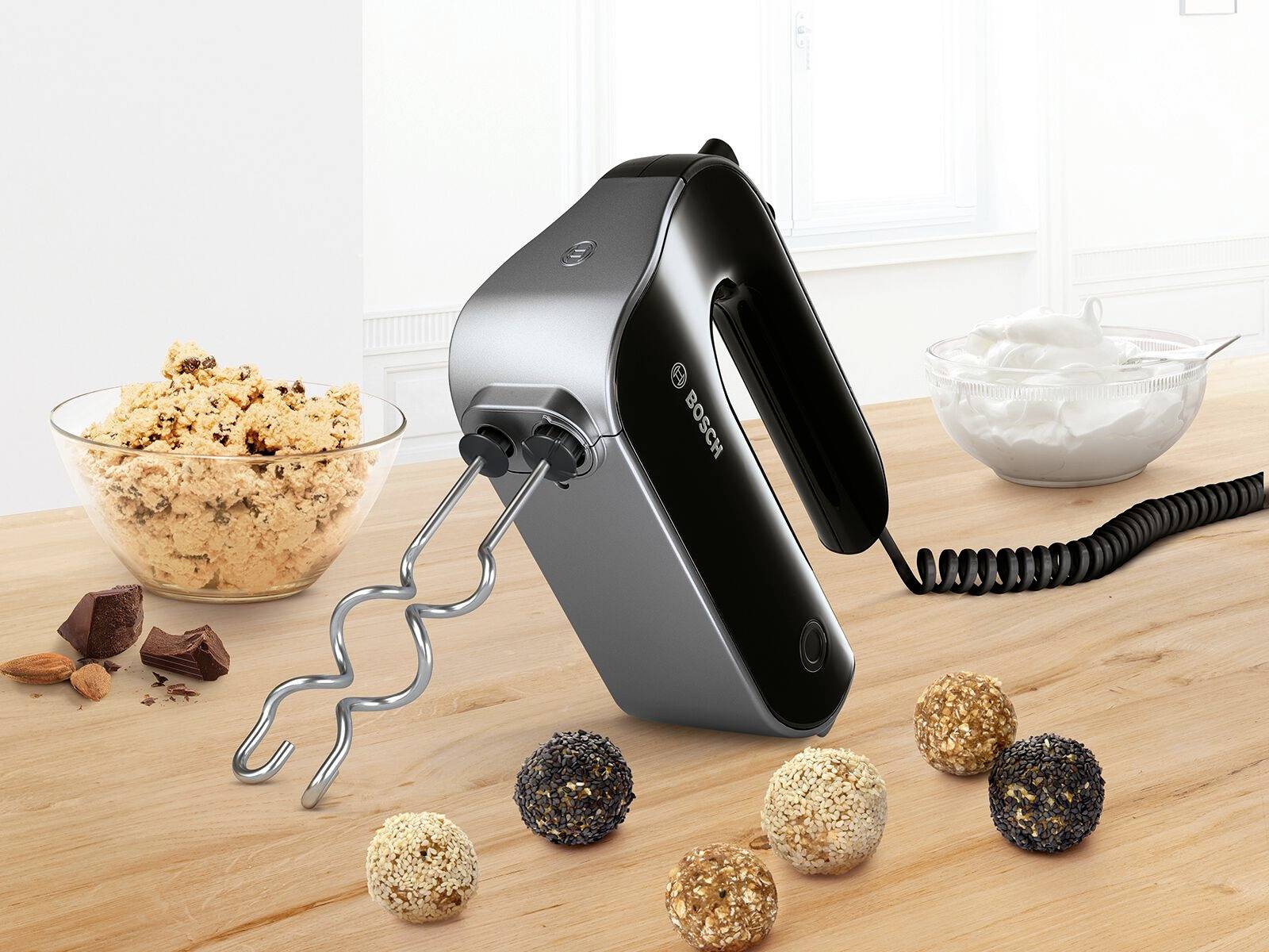 Schwarzer Handmixer auf Holztisch, umgeben von Keksteig und Schlagsahne. Mixstäbe sind aufgesetzt, Kabel im Hintergrund.