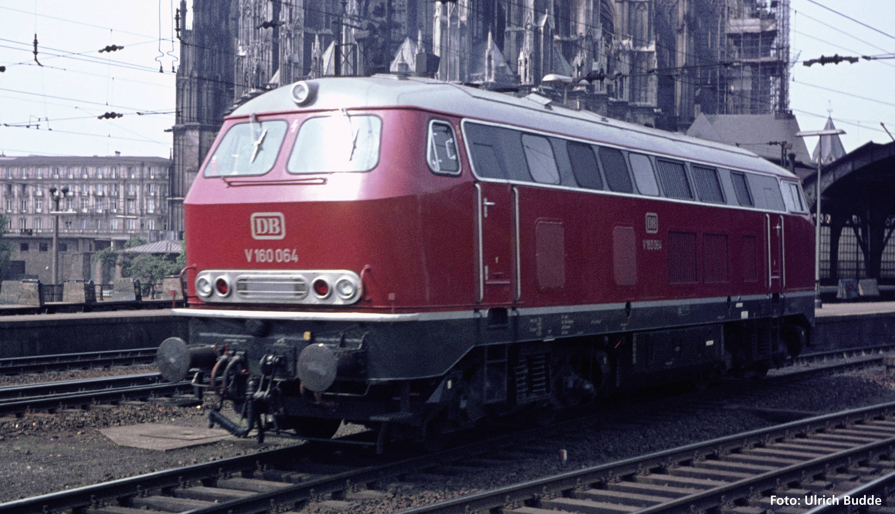 Eine rote Diesellok der Deutschen Bahn steht auf Schienen vor einem alten, großen Gebäude. Im Hintergrund sind weitere Schienen und Gebäude zu sehen.