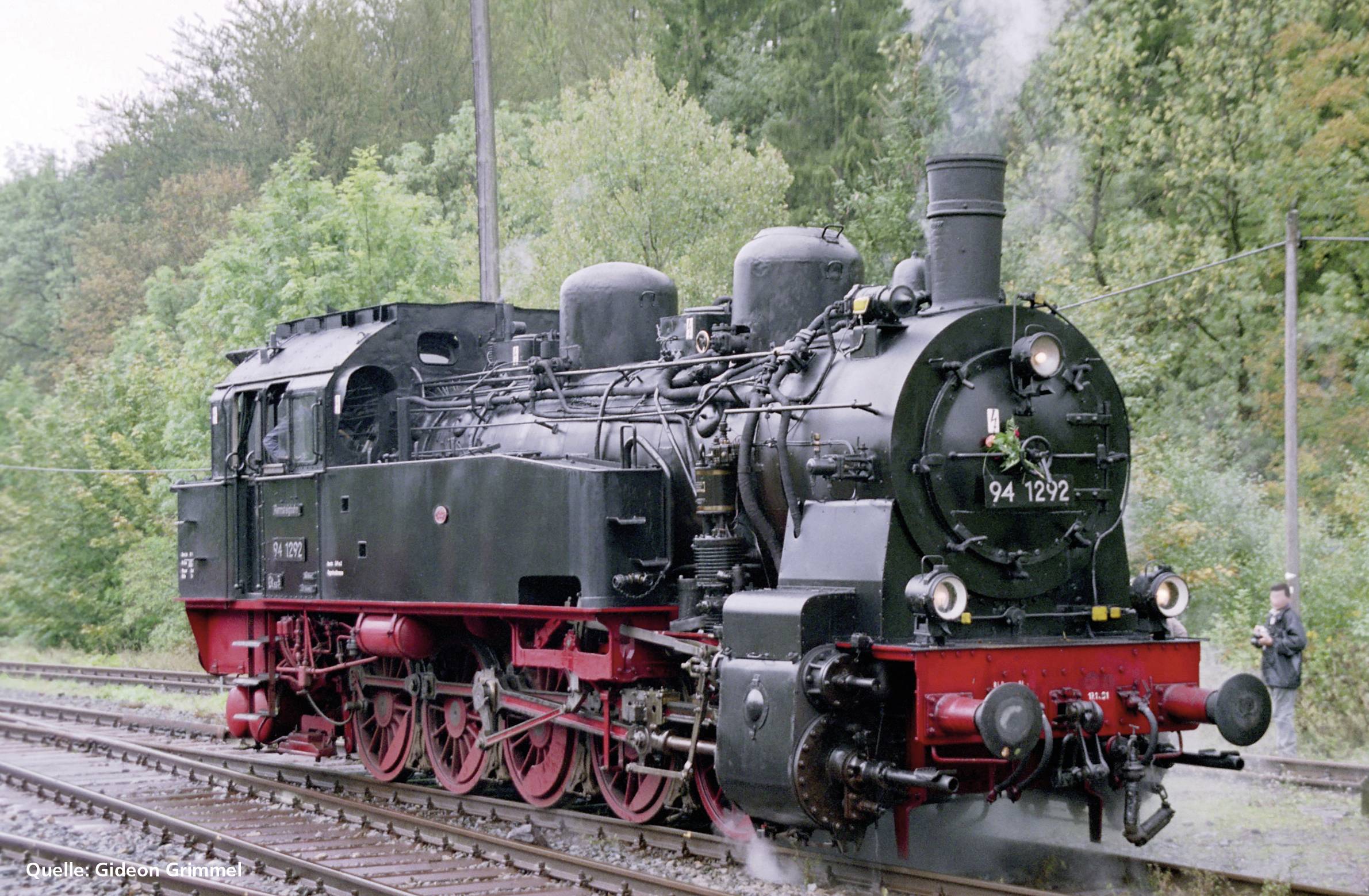Eine schwarze Dampflokomotive auf einem Bahngleis, umgeben von Bäumen im Hintergrund, mit leichtem Rauch aus dem Schornstein.