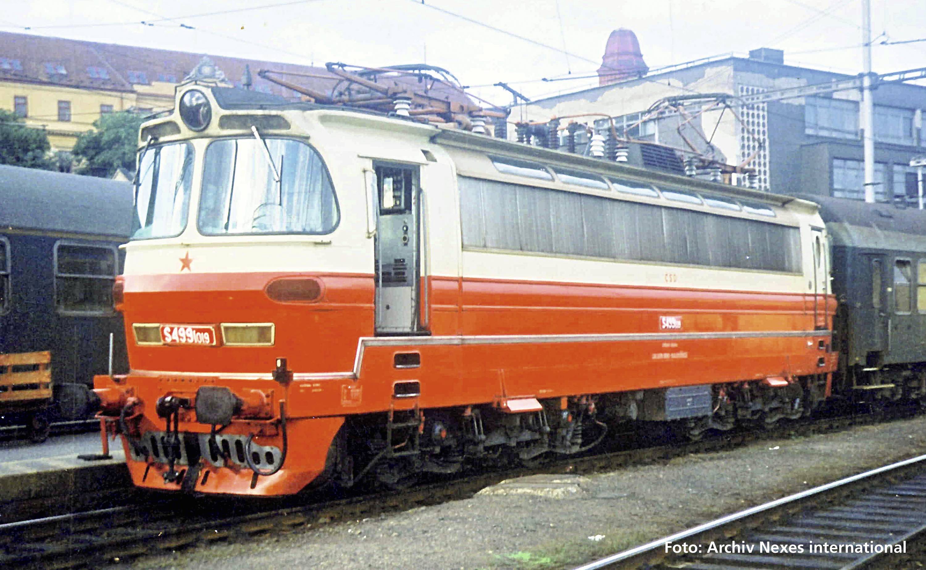 Eine rot-weiße Elektrolokomotive steht auf einem Bahnhofsgleis. Sie hat runde Fenster an der Vorderseite und ein auffälliges Sternsymbol.