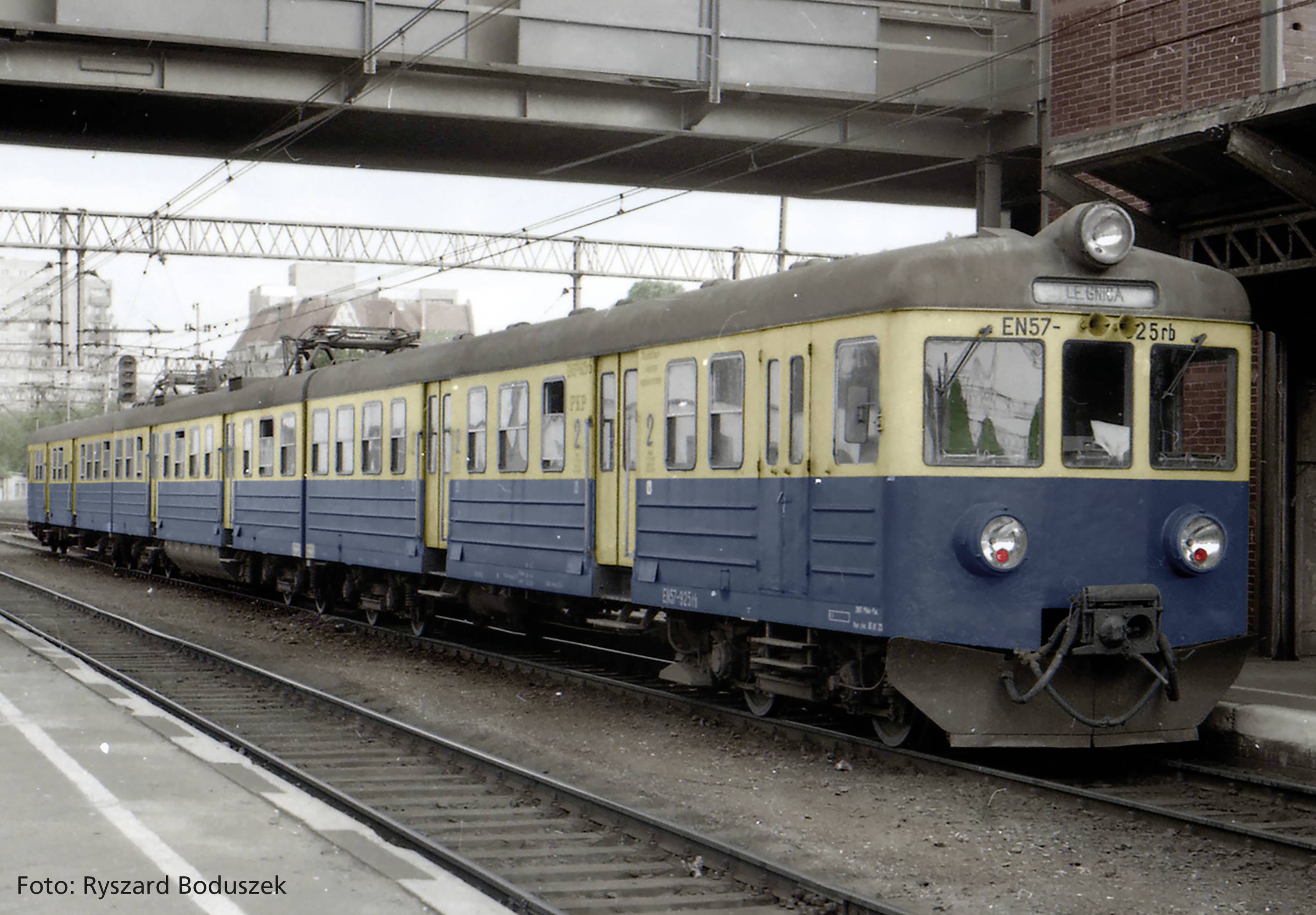 Ein alter, blauer und gelber Zug steht auf einem Bahngleis in einem städtischen Bahnhof. Hintergrund zeigt Industriegebäude und Stromleitungen.