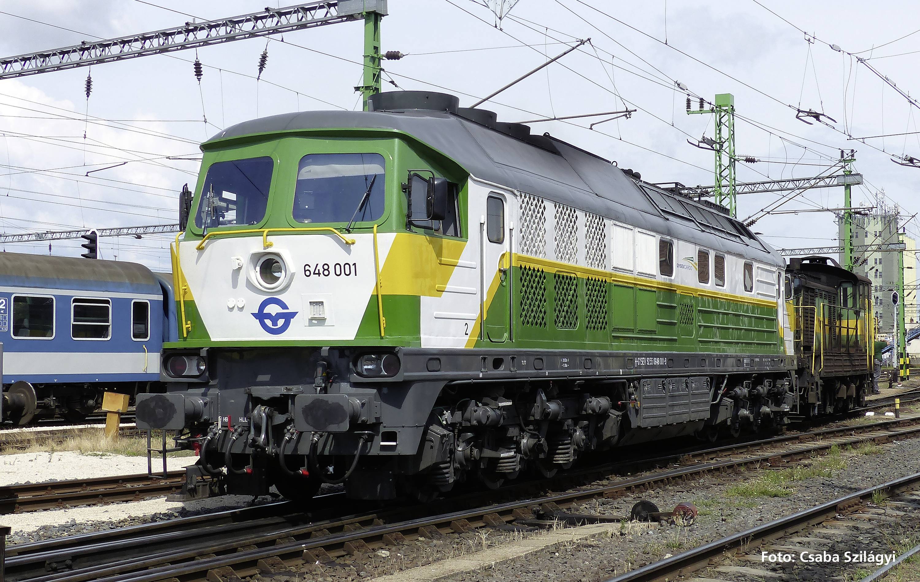 Ein grüner und weißer Zug mit der Nummer '648 001' auf einem Bahngleis, umgeben von Oberleitungen und weiteren Zügen im Hintergrund.