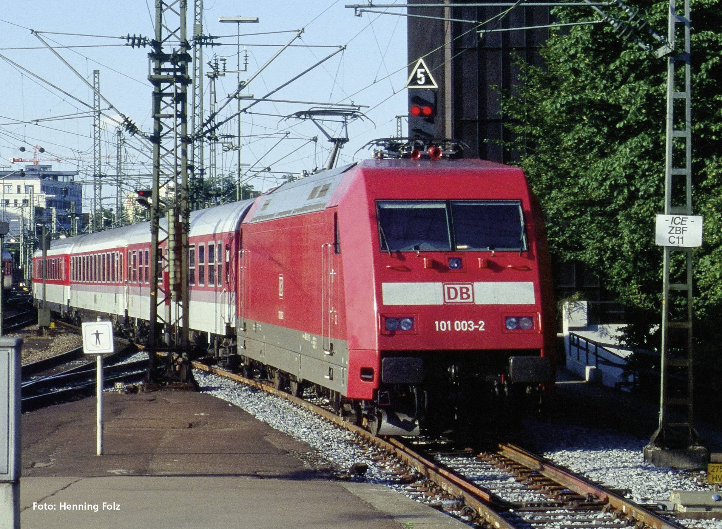 Ein roter Zug der Deutschen Bahn fährt auf einem Gleis durch eine städtische Gegend. Oberleitungen und Signale sind sichtbar.