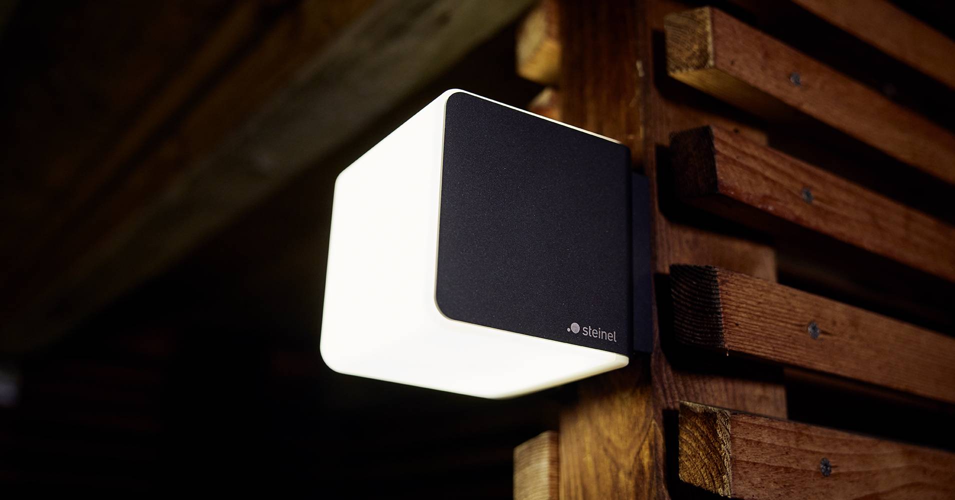 Wandleuchte mit modernem, kubischem Design, leuchtet warmweiß und ist an einer hölzernen Wand montiert.