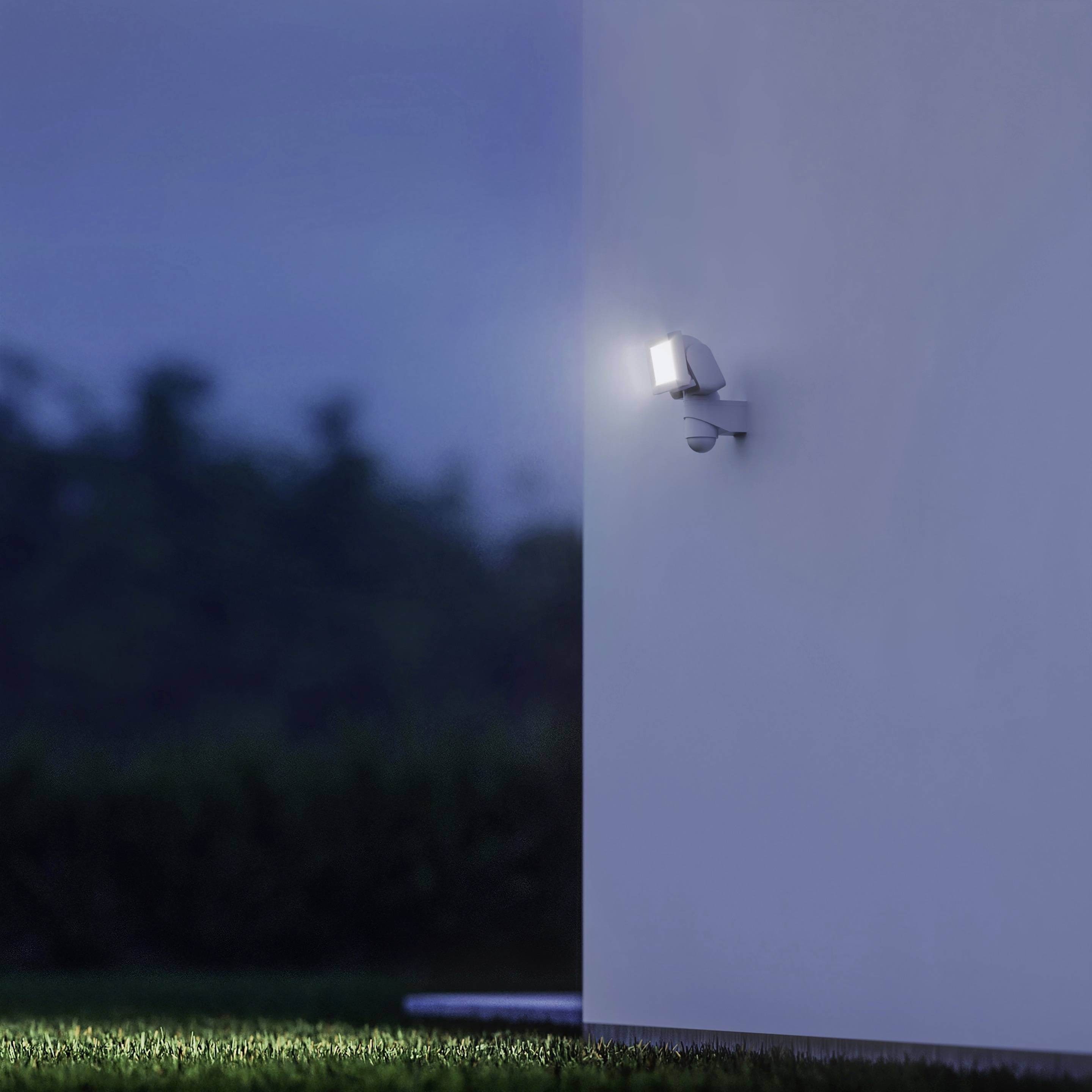 Eine Außenlampe leuchtet an einer Wand bei Dämmerung oder Nacht. Im Hintergrund ist eine unscharfe, dunkle Landschaft zu sehen.