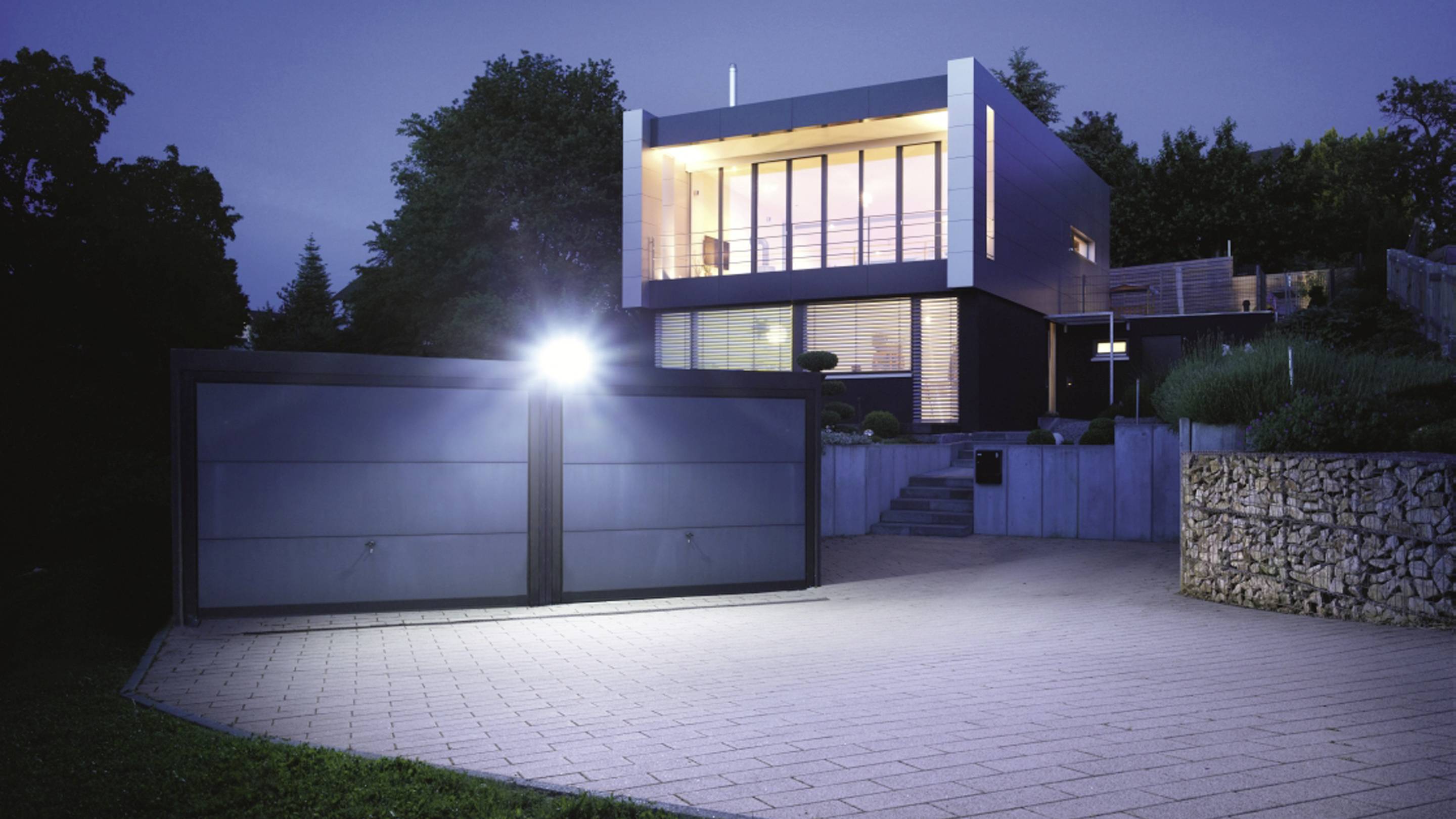 Moderne zweistöckige Villa mit großer Glasfassade, beleuchtet bei Nacht. Vor dem Haus eine breite Einfahrt mit geschlossenem Tor.