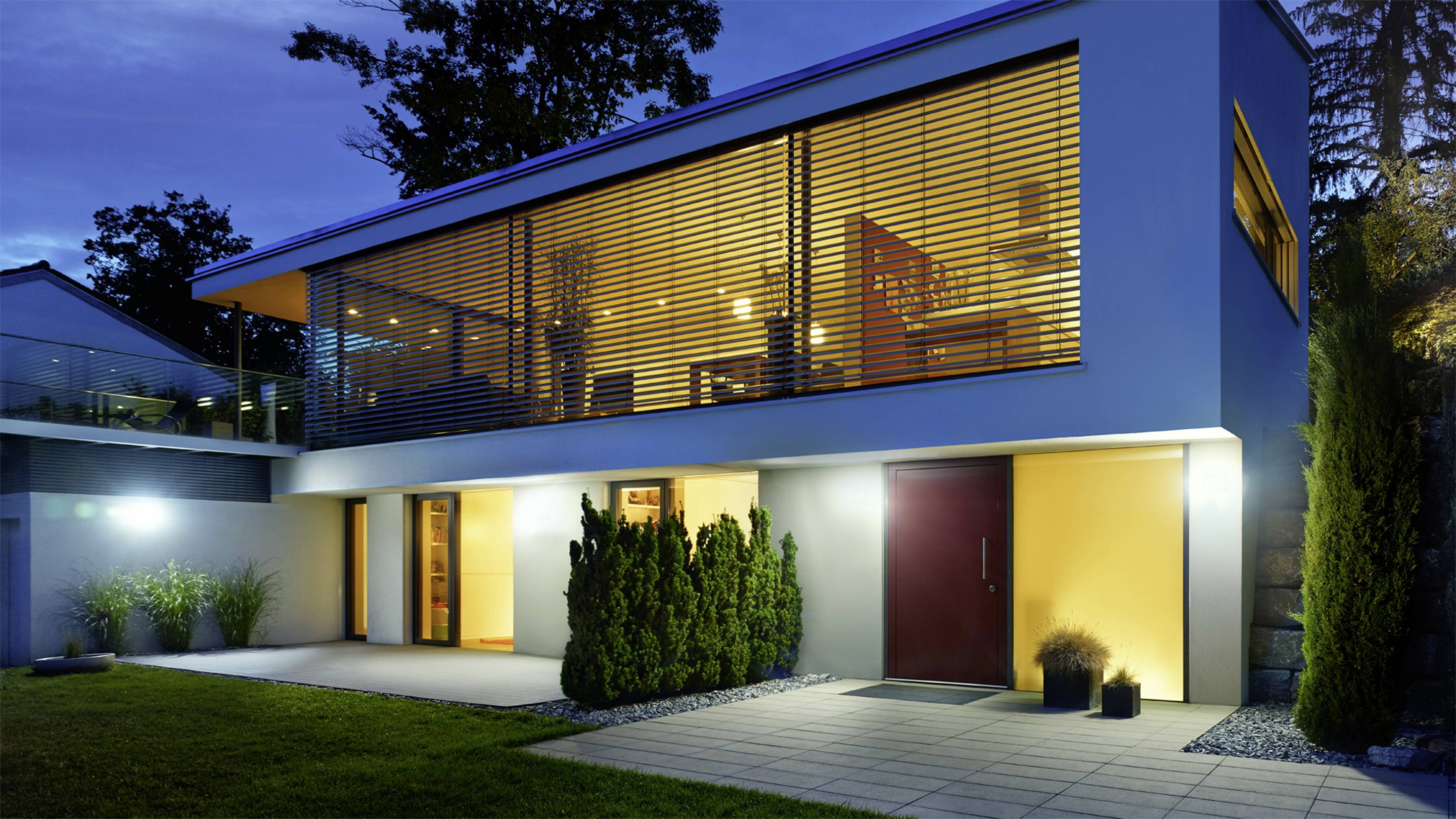 Modernes zweistöckiges Haus bei Abenddämmerung, mit großer verglaster Fassade im Obergeschoss, beleuchteter Terrasse und gepflegtem Garten.