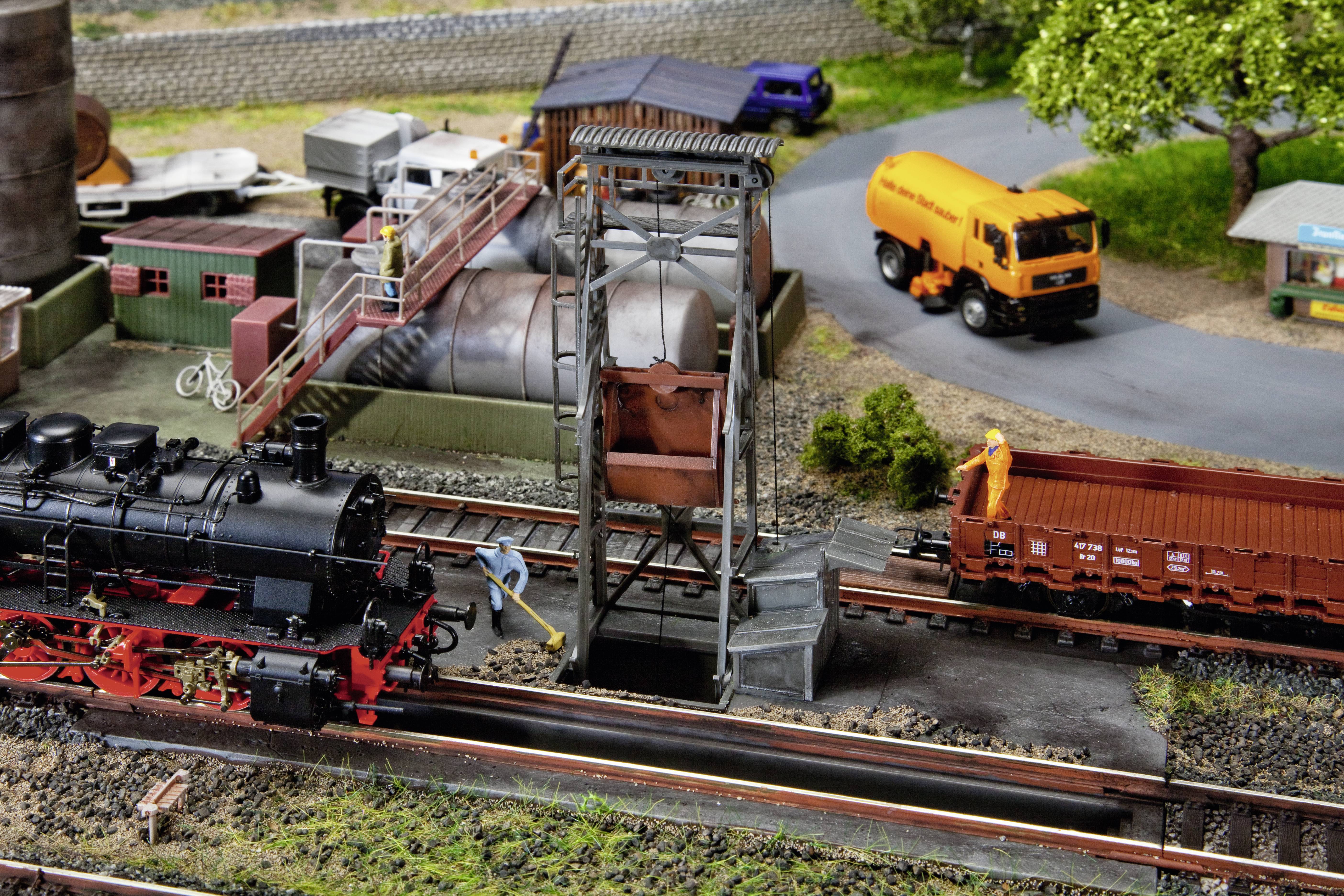 Modelleisenbahnszene mit einer Dampflokomotive, die neben einem Kran hält. Ein LKW und Güterwaggons sind im Hintergrund sichtbar.