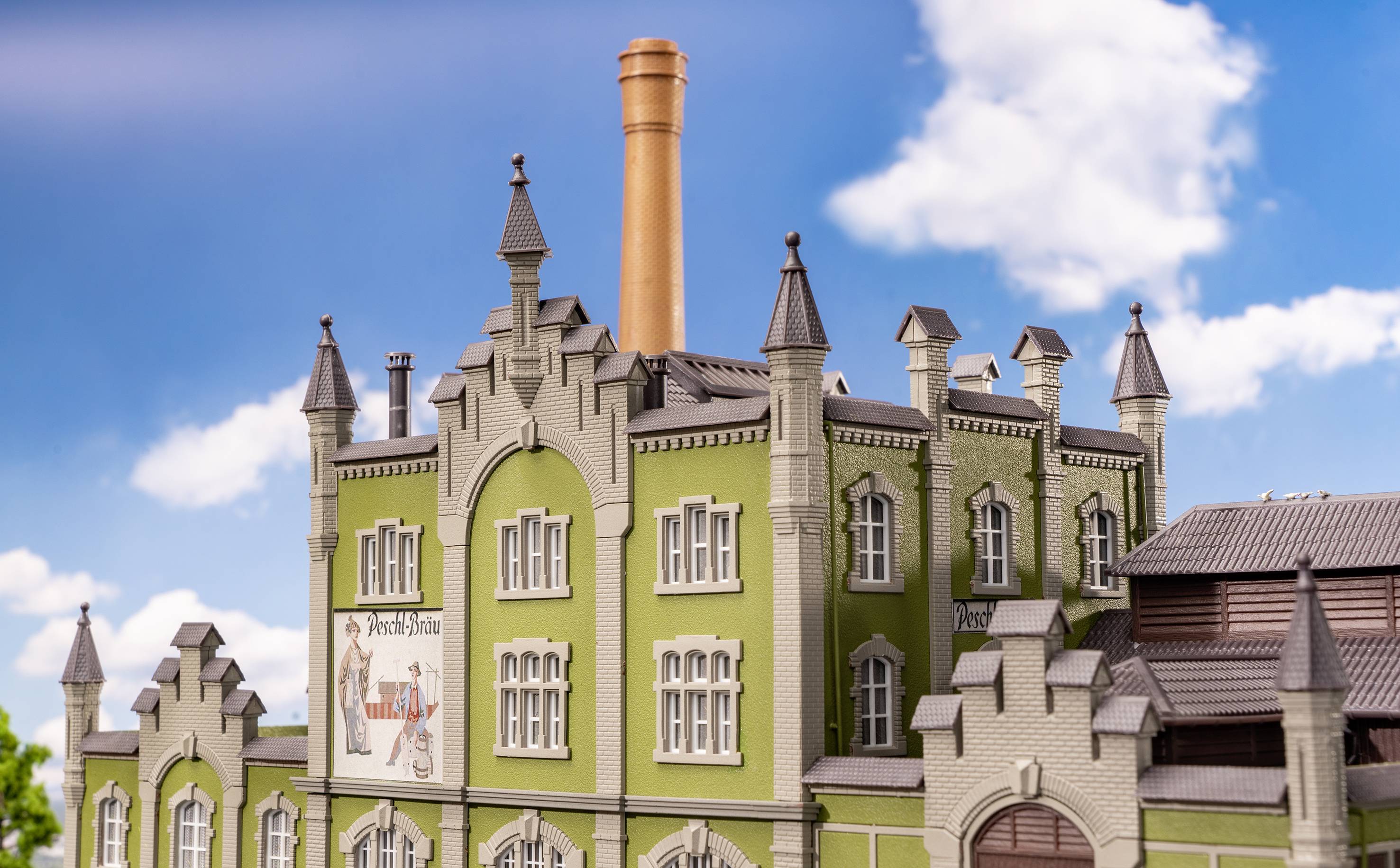 Ein Modell einer alten Fabrik mit grüner Fassade und Ziegelturm vor blauem Himmel. Plakat zeigt 'Brauerei'.