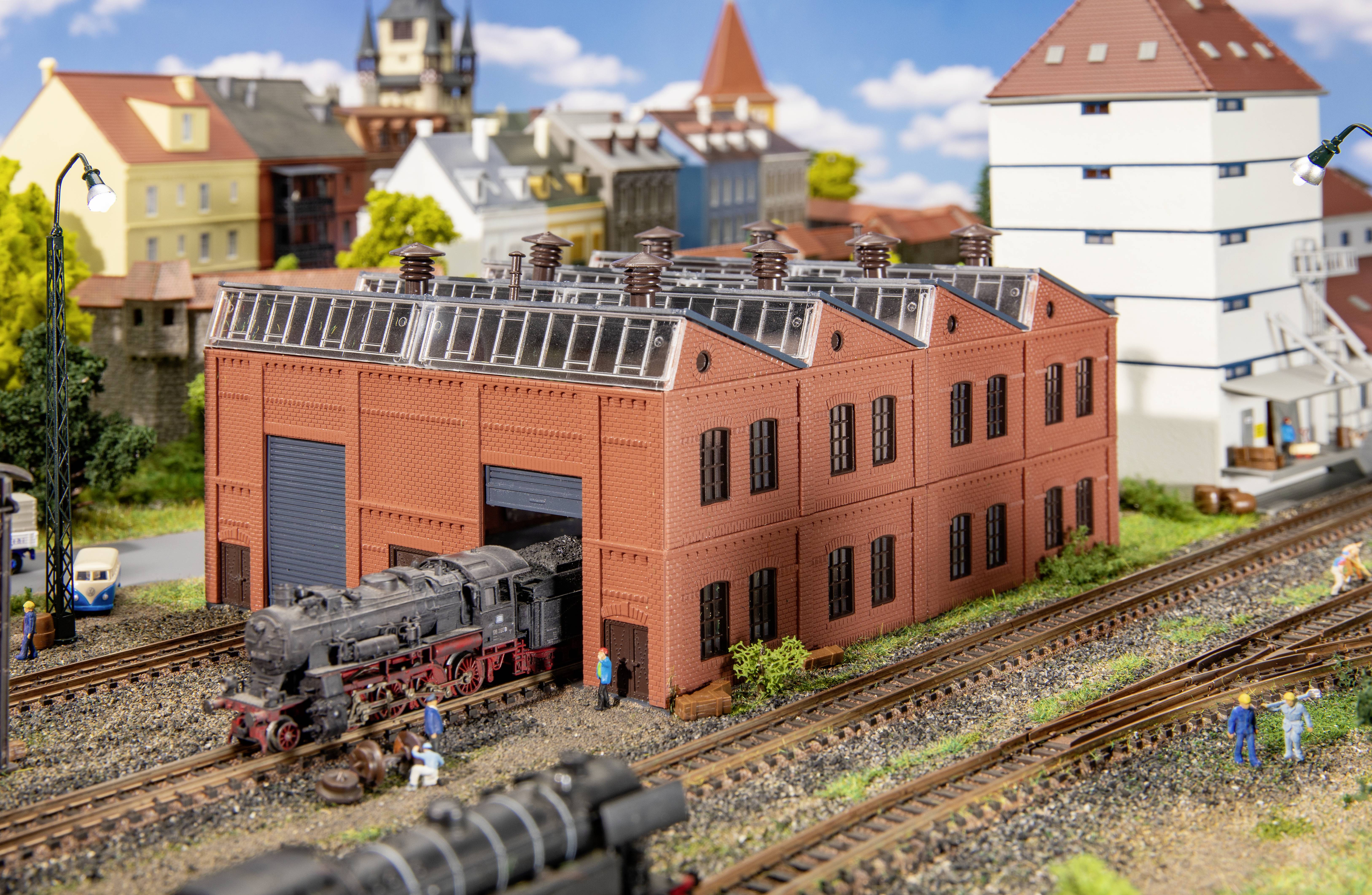 Ein Modellbahndiorama zeigt eine schwarze Dampflokomotive, die aus einem roten Backsteingebäude fährt. Im Hintergrund sind Gebäude und Bäume zu sehen.