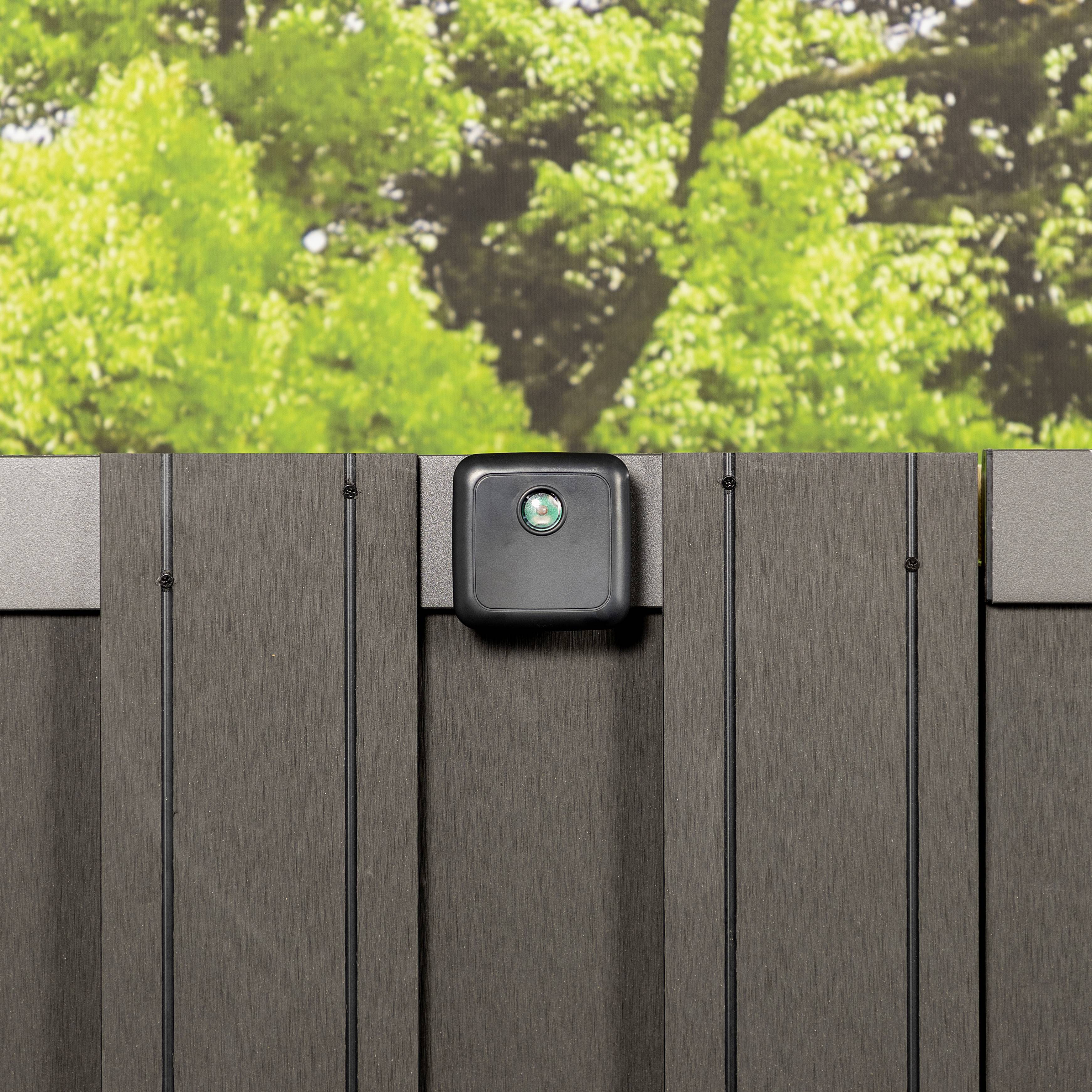 Ein schwarzes Außengerät, möglicherweise eine Kamera oder Licht, ist an einer vertikalen, grauen Zaunplatte befestigt; im Hintergrund Bäume.