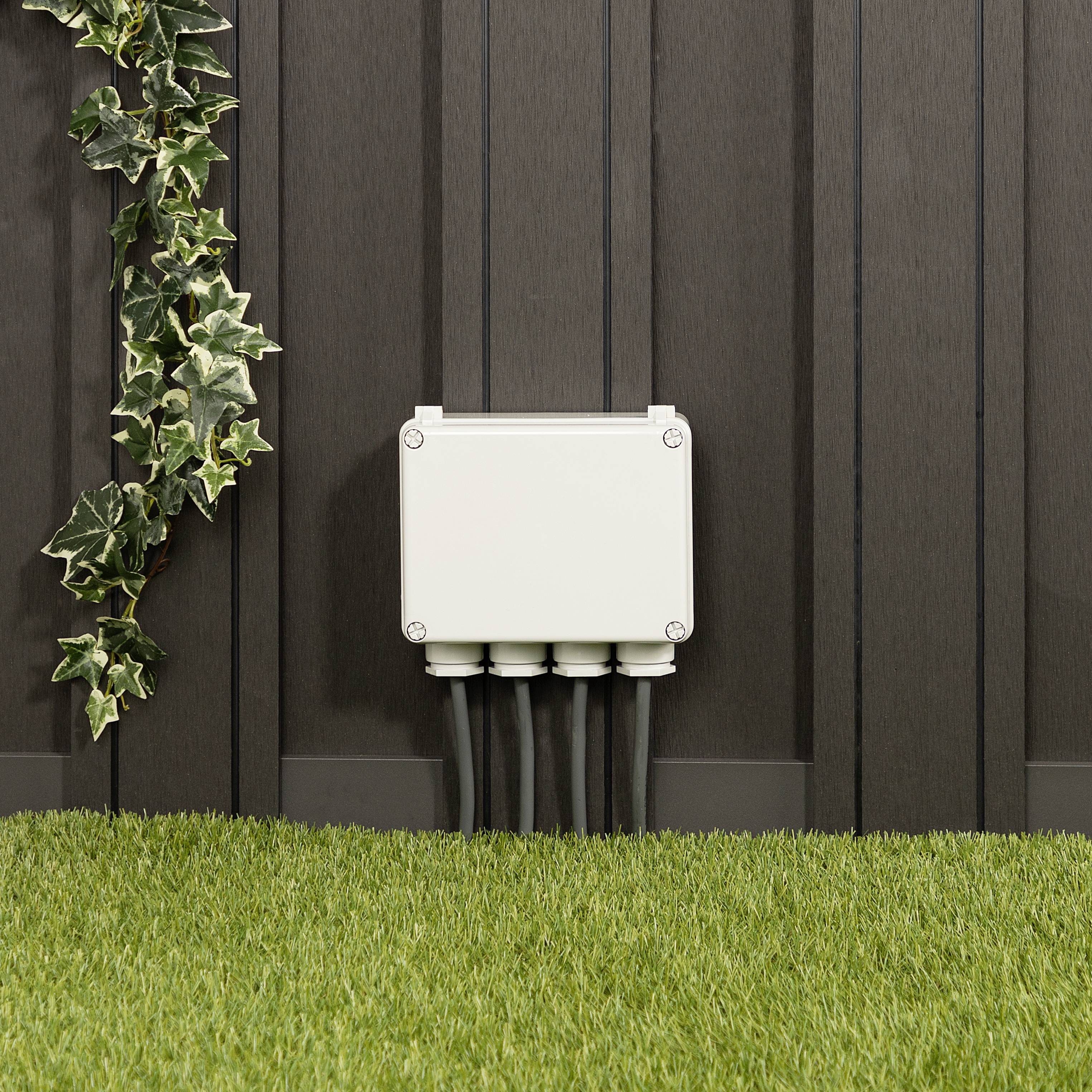 Ein elektrischer Verteilerkasten ist an einer Holzwand montiert. Kabel führen nach unten. Links schlängeln sich grüne Efeuranken.
