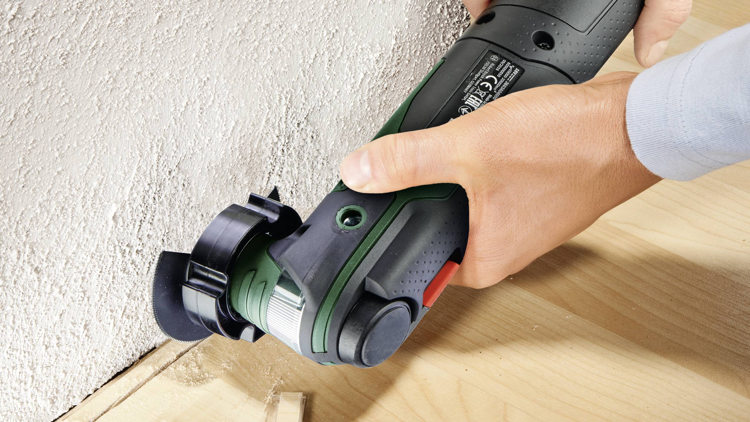 Eine Person verwendet ein elektrisches Multifunktionswerkzeug, um ein Holzstück nahe einer Wand präzise zu schneiden.