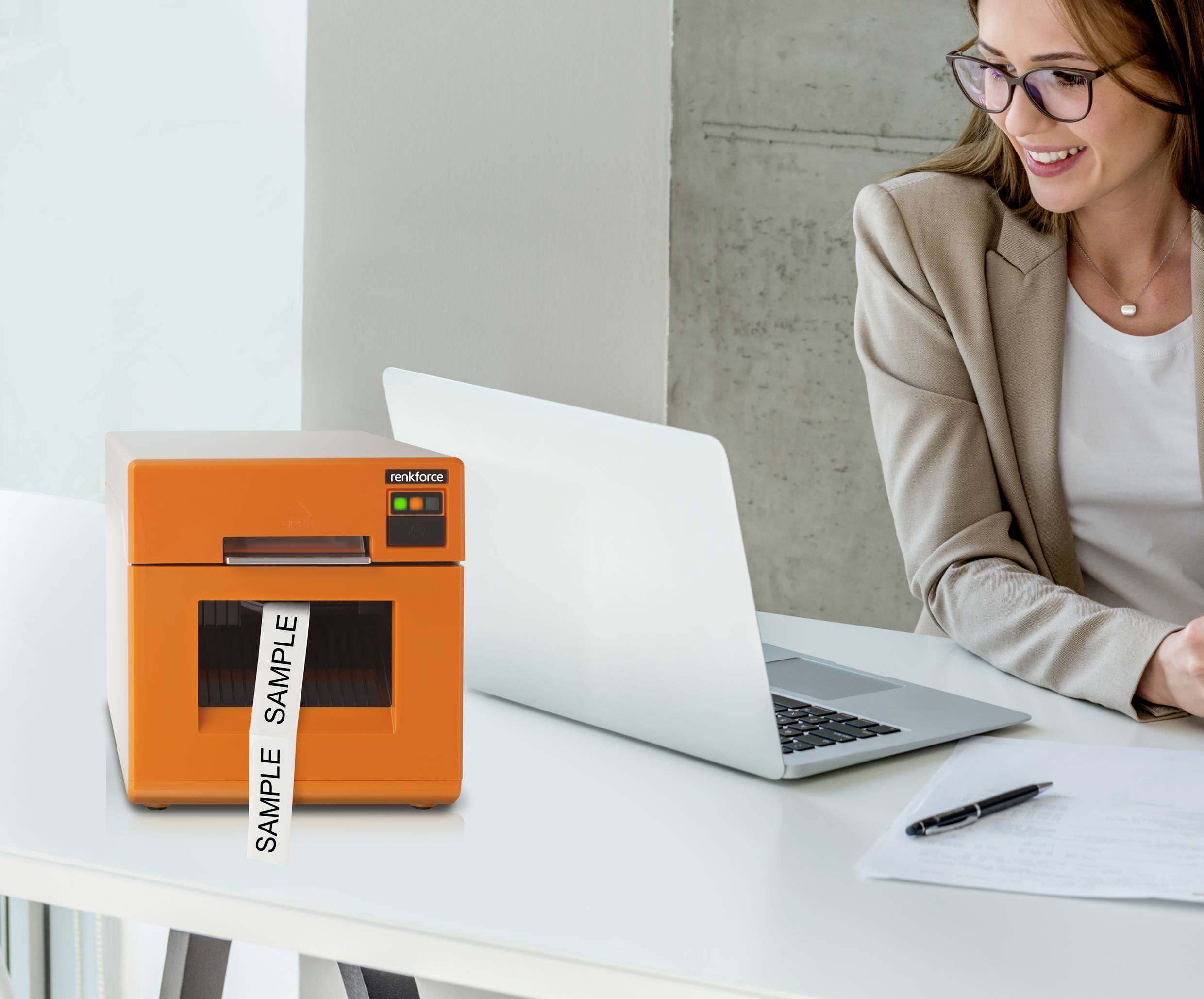 Frau sitzt an einem Schreibtisch mit Laptop, Block und Stift. Ein orangefarbener Drucker mit 'SAMPLE'-Etikett steht daneben.
