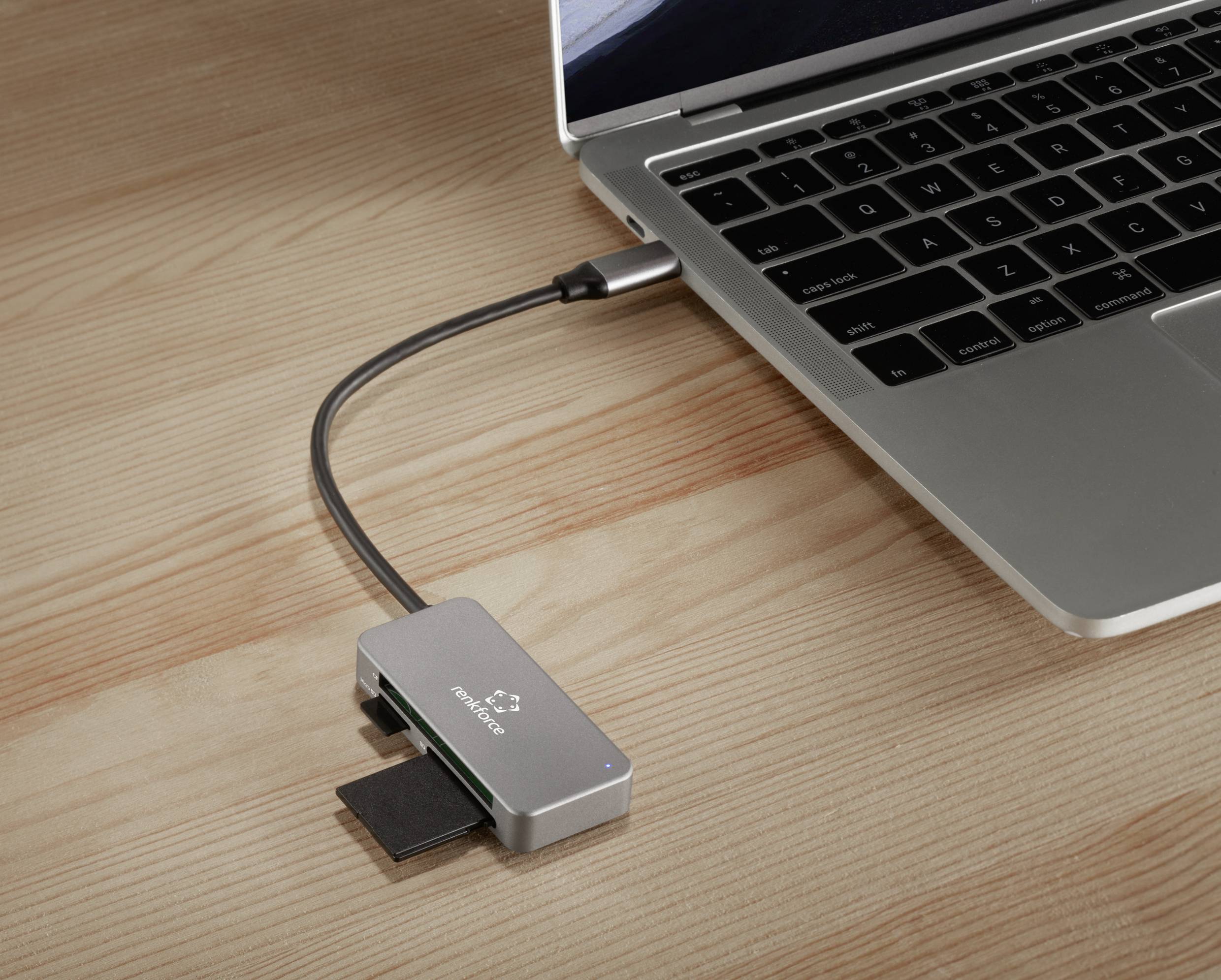 Ein Laptop ist über ein USB-C-Hub mit mehreren Anschlüssen verbunden, darunter für SD-Karten. Das Setup liegt auf einem Holztisch.
