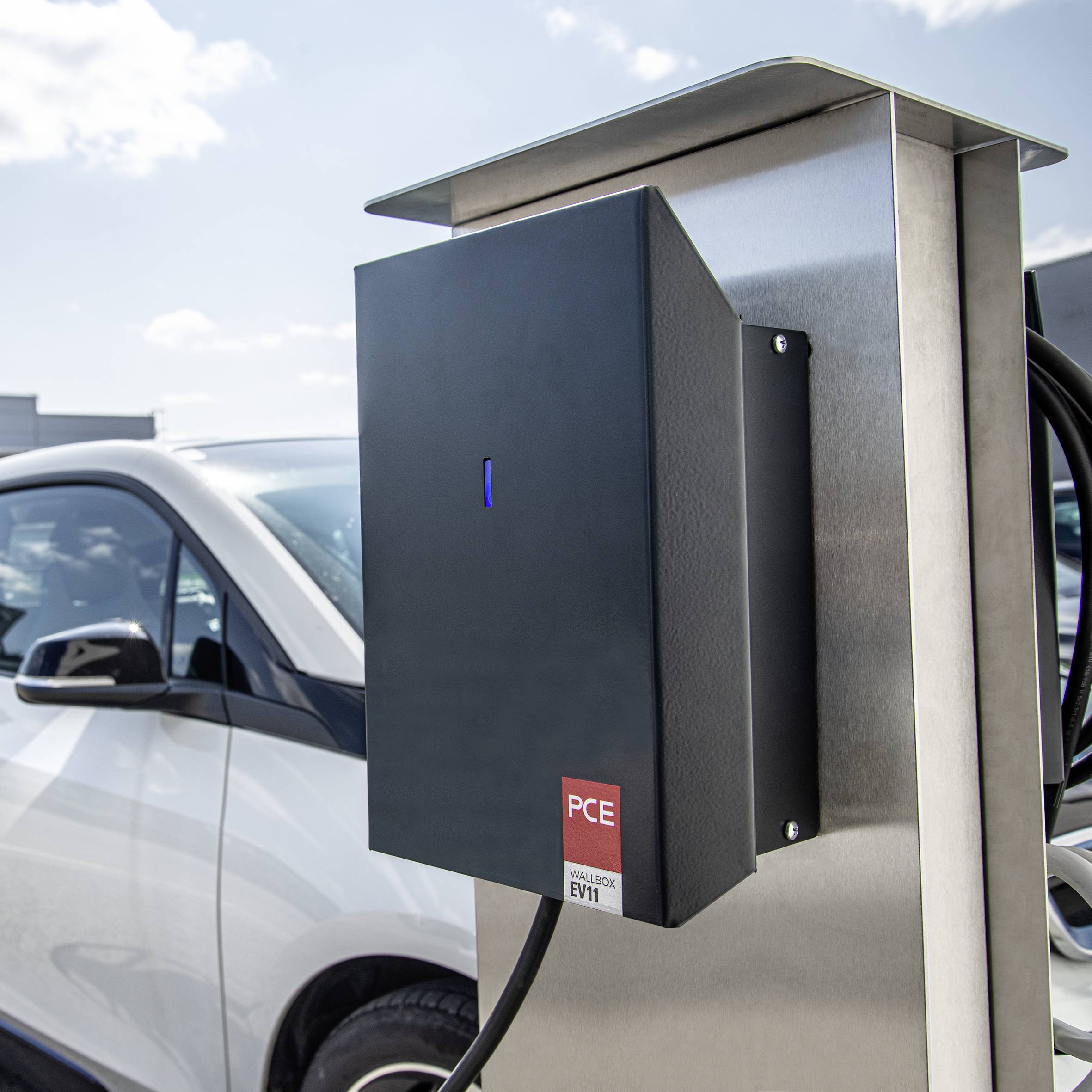 Ein Elektroauto wird an einer Ladestation auf einem Parkplatz aufgeladen. Im Hintergrund sind weitere Autos und Gebäude sichtbar.