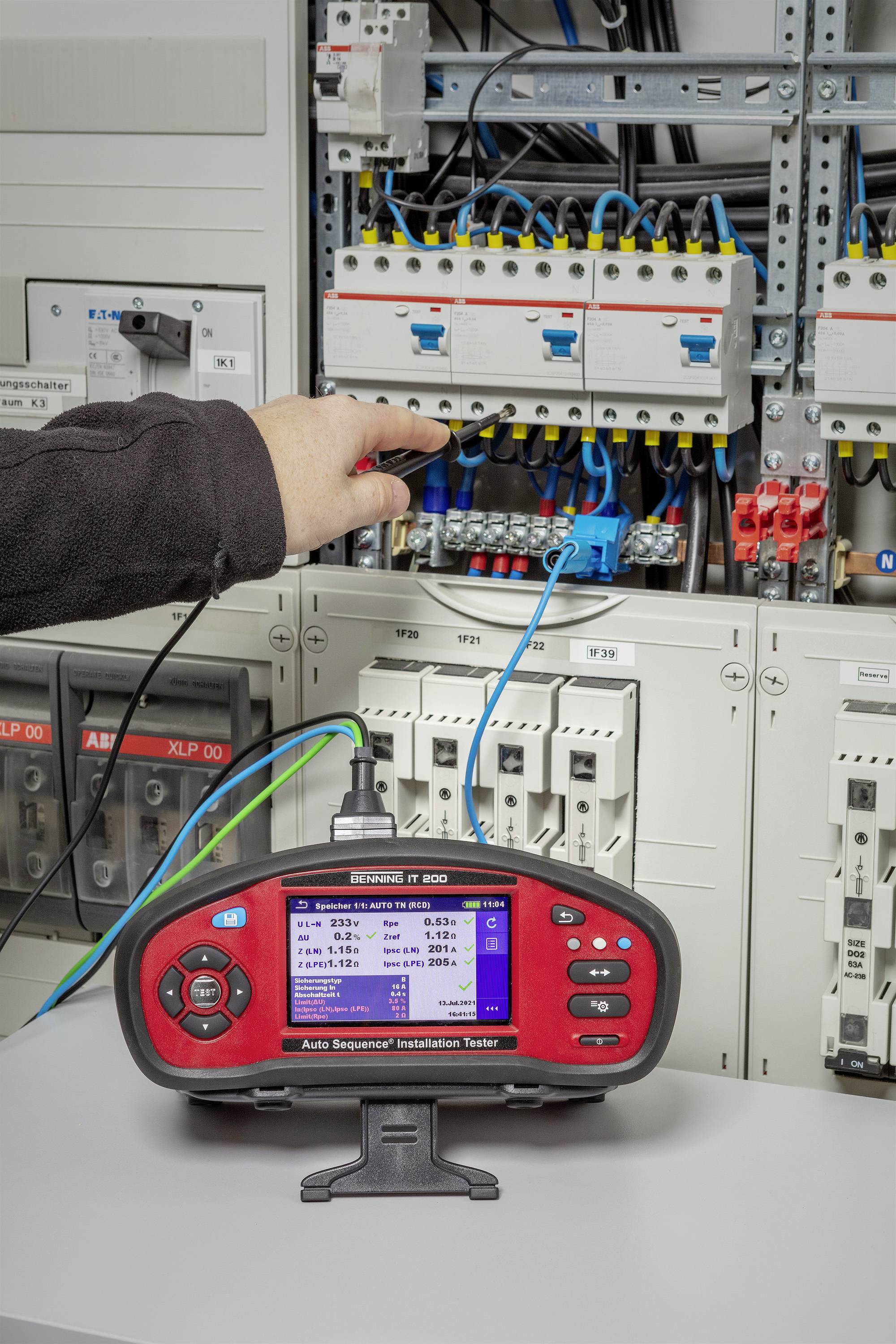 Eine Person testet ein elektrisches Schaltsystem mit einem Prüfgerät. Das Gerät zeigt Ergebnisse auf einem Display an. Kabel sind verbunden.