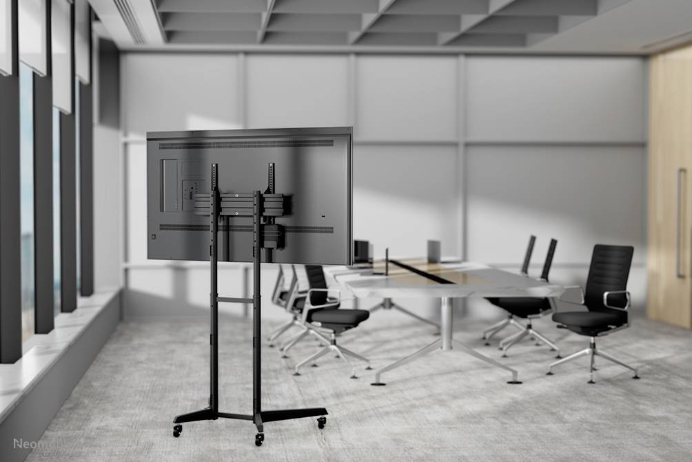 Moderner Konferenzraum mit einem großen Bildschirm auf einem Ständer, Tisch und schwarzen Stühlen. Großes Fenster sorgt für natürliches Licht.