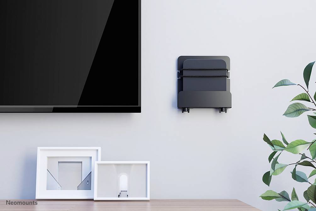 Flachbildfernseher an der Wand neben dekorativem Wandregal mit zwei Bilderrahmen, einer leer, einer mit Kunst. Pflanze rechts im Bild.