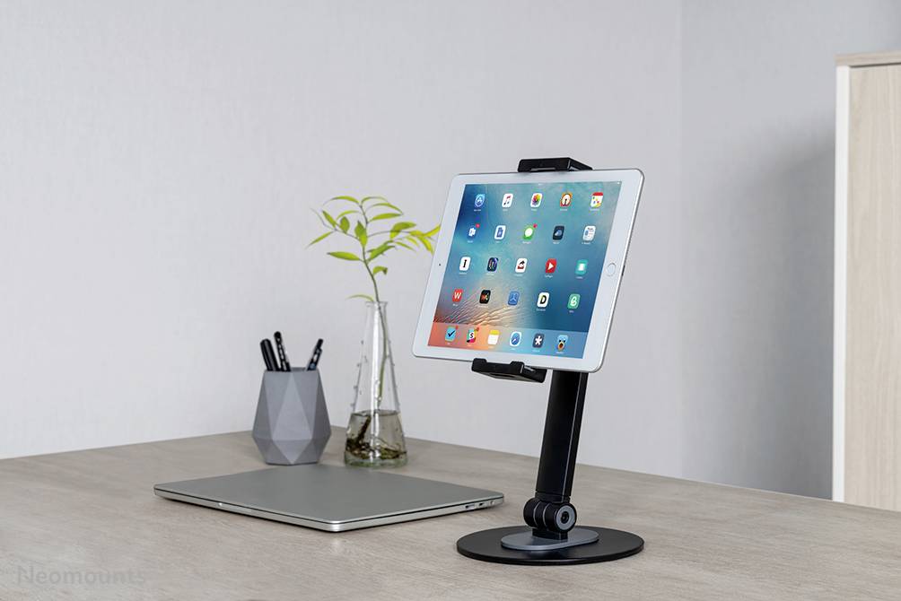 Ein Tablet auf einem schwarzen Ständer, platziert auf einem Schreibtisch neben einem geschlossenen Laptop, einer Vase mit Pflanzen und einem Stiftbehälter.